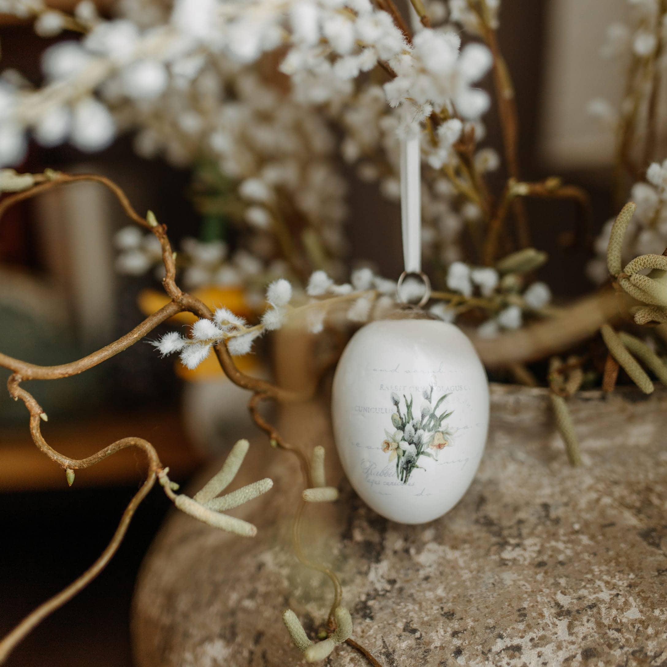 Adorno de Pascua de cerámica blanca en forma de huevo con motivo floral colgado de una rama decorativa