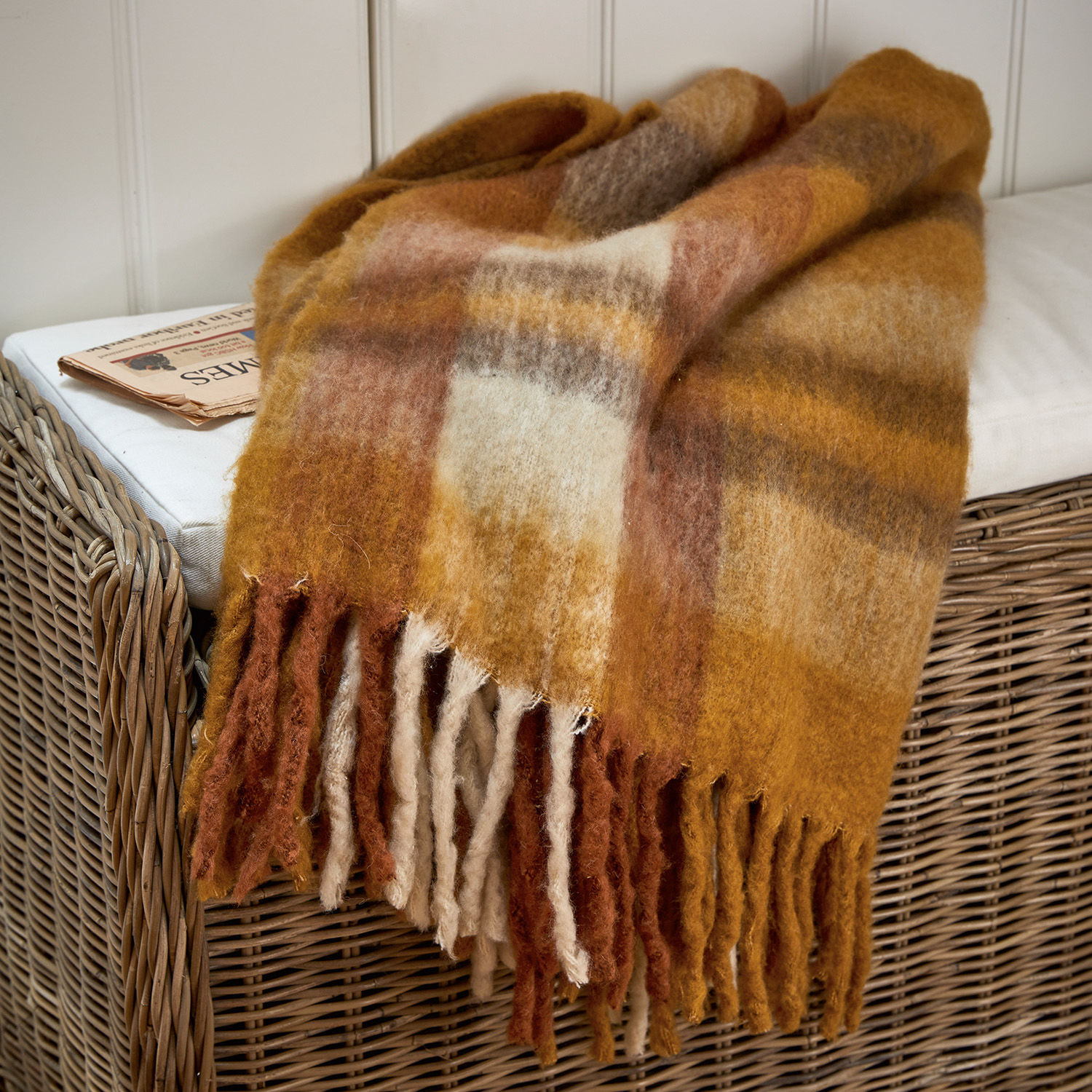 Plaid Alborg Manta de lana suave en tonos cálidos marrón y beige con flecos sobre sillón de mimbre