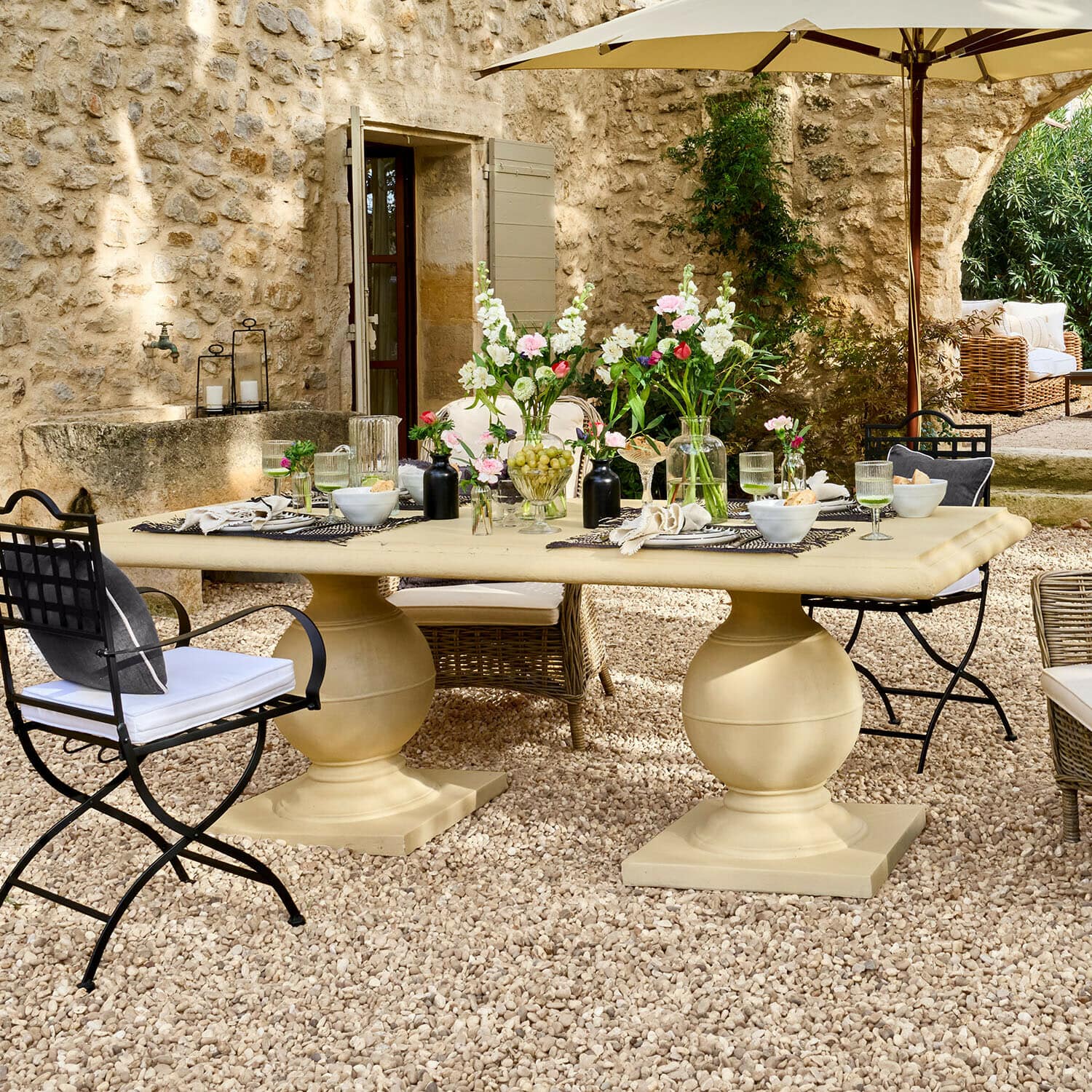 Mesa Talvero Gran mesa de jardín beige de piedra en patio de grava con flores y sillas de metal y mimbre
