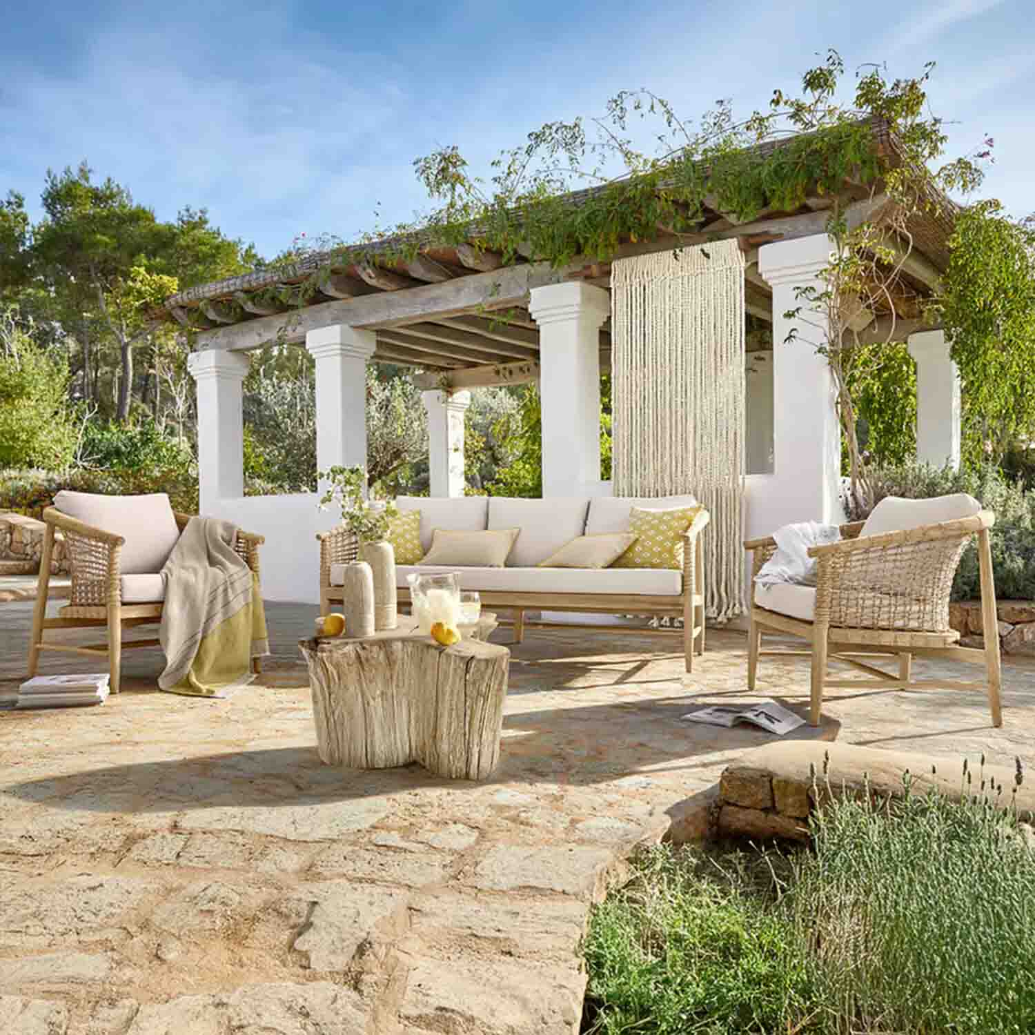Conjunto de muebles de jardín de madera clara con cojines crema en terraza de piedra bajo pérgola con plantas