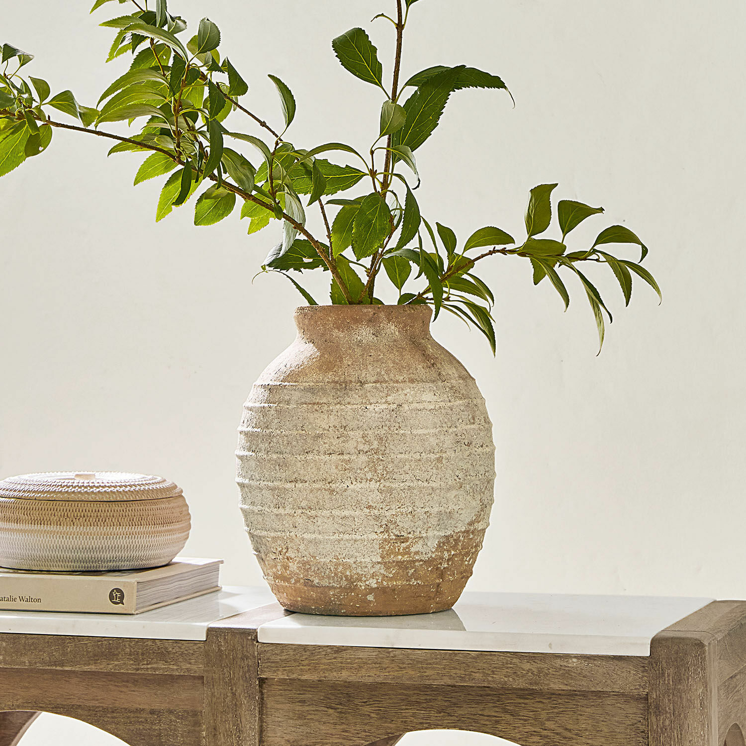 Jarrón rústico de cerámica beige y marrón con ramas verdes sobre mesa de madera