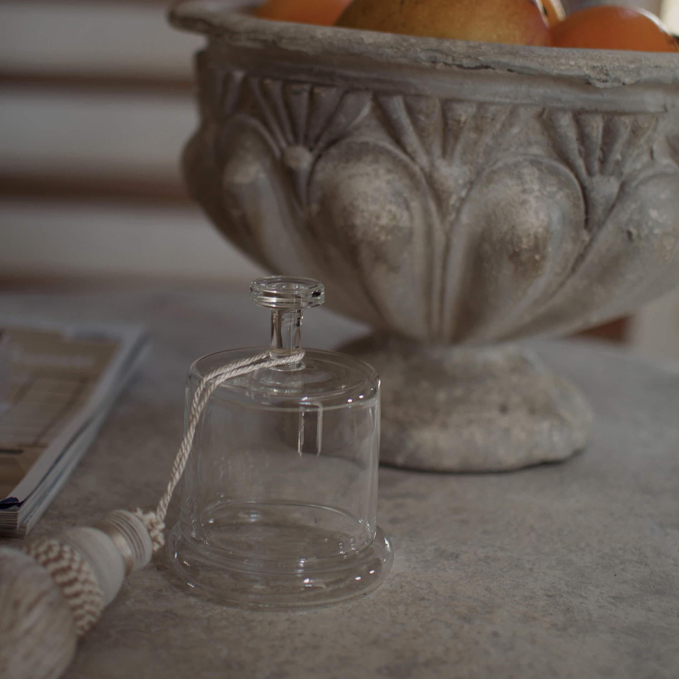 Campana decorativa de vidrio transparente con cuerda blanca sobre mesa de piedra junto a un cuenco ornamentado
