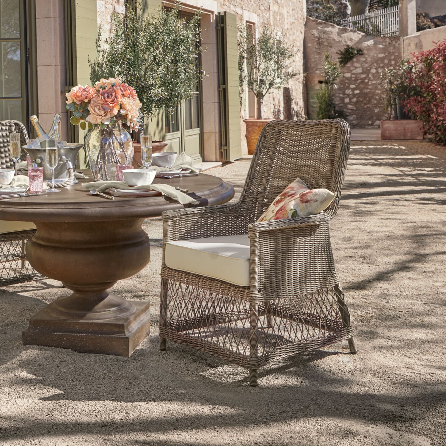 Sillón Fenris Silla de ratán gris con cojín crema y cojín floral junto a mesa redonda en exterior