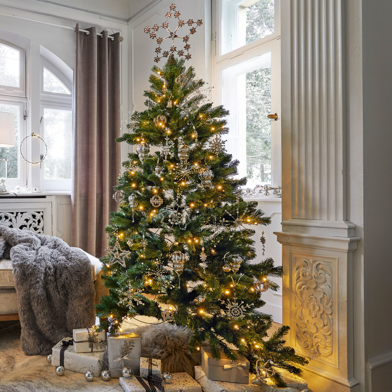Set de adornos navideños Bjouterie Árbol de Navidad verde con adornos plateados y luces en sala luminosa con regalos debajo