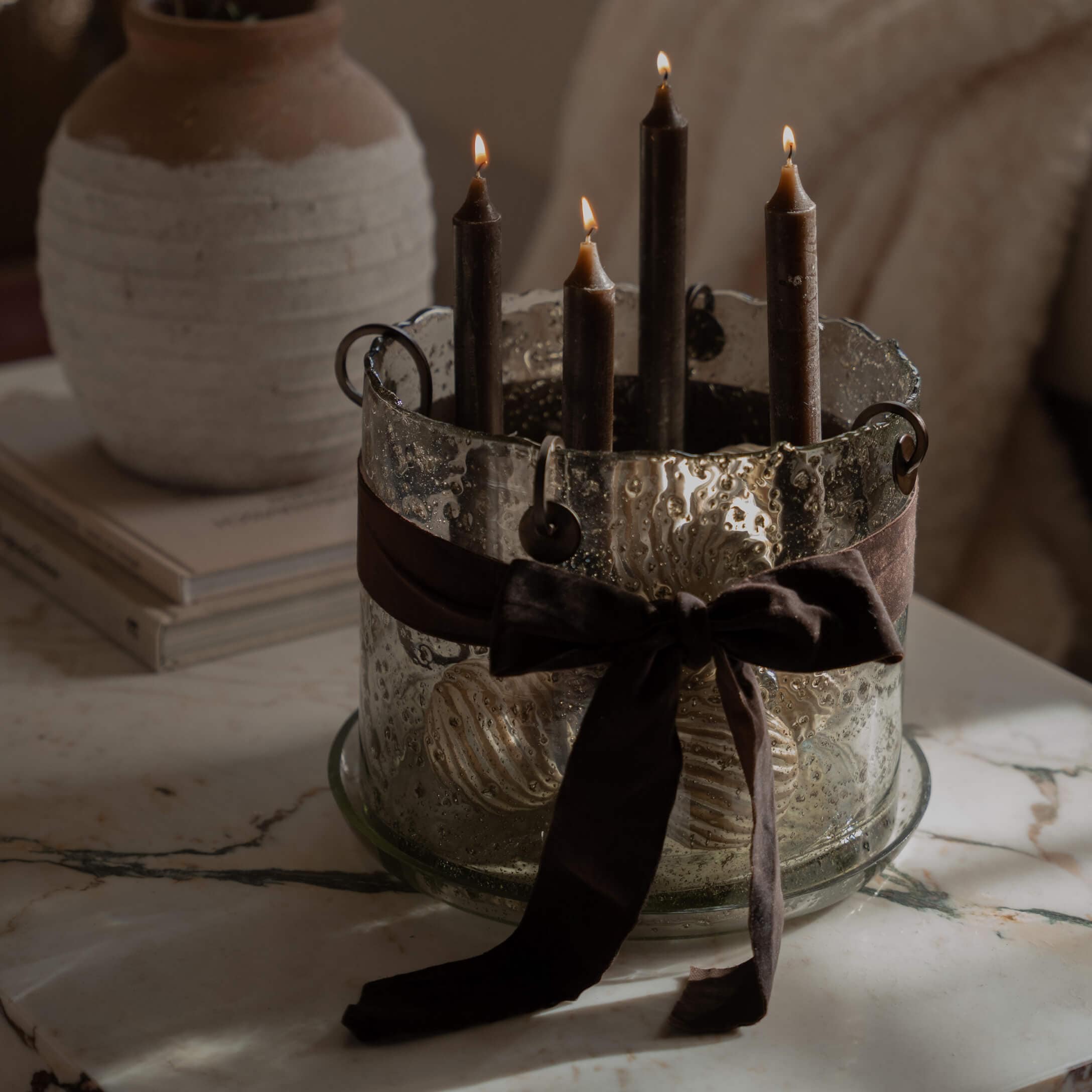 Portavelas de vidrio con cuatro velas negras y lazo marrón sobre mesa de mármol