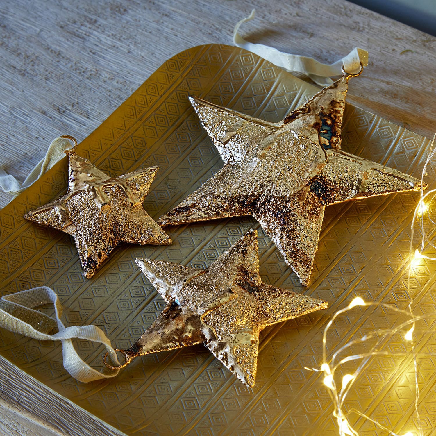 Set de 3 colgantes Myrska Conjunto de tres estrellas decorativas de metal dorado sobre bandeja dorada con luces LED