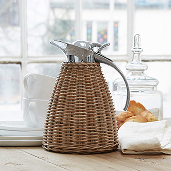 Jarra de ratán con tapa metálica sobre mesa de madera, ideal para servir bebidas en estilo rústico
