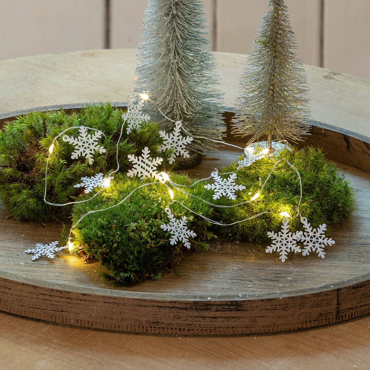 Luces de hadas Azmira Guirnalda decorativa con copos de nieve blancos y luces LED sobre musgo verde en bandeja de madera