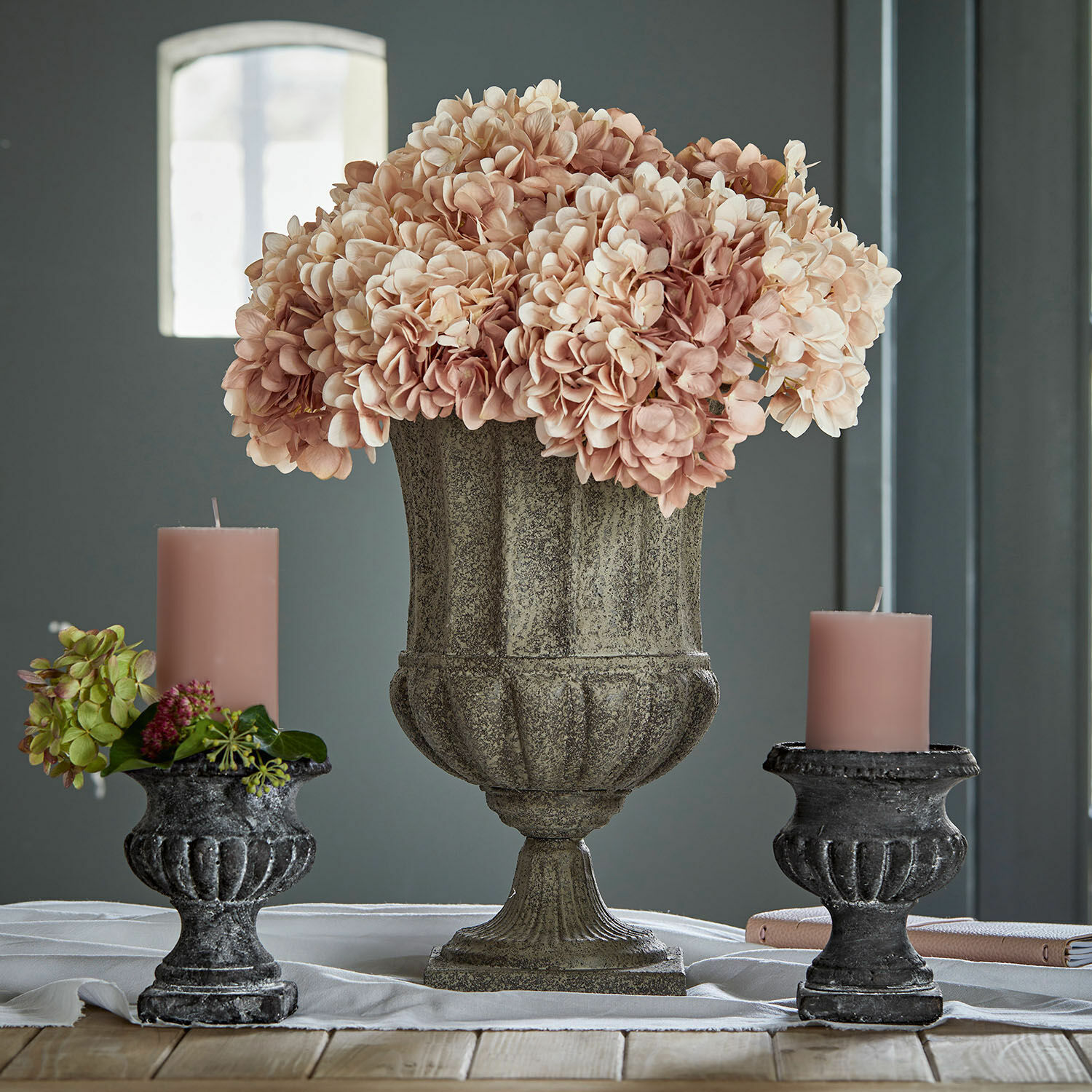Ánfora Renny Jarrón decorativo de piedra con flores rosas y dos candelabros negros con velas rosas sobre mesa de madera
