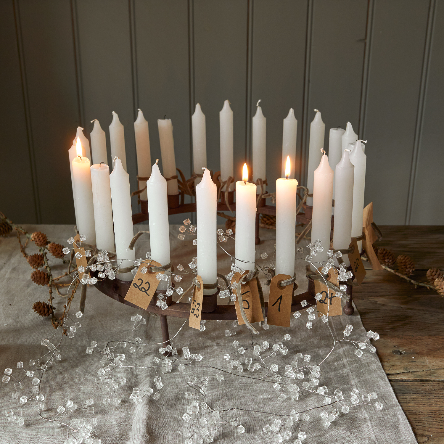 Corona de adviento metálica con 24 velas blancas numeradas y etiquetas de papel sobre mantel de lino