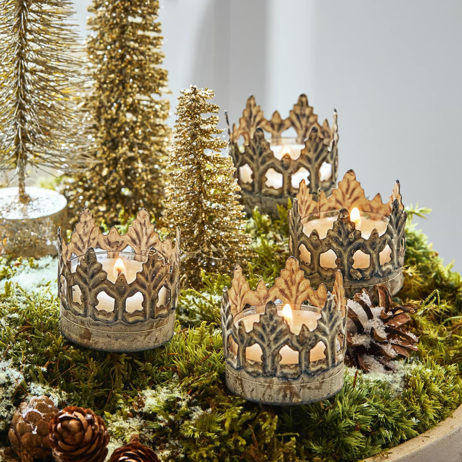 Portavelas metálicos en forma de corona sobre musgo verde con decoración navideña y árboles dorados