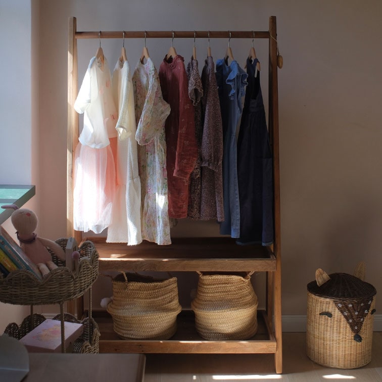 Perchero de madera con vestidos en tonos pastel y cestas de mimbre en habitación infantil luminosa
