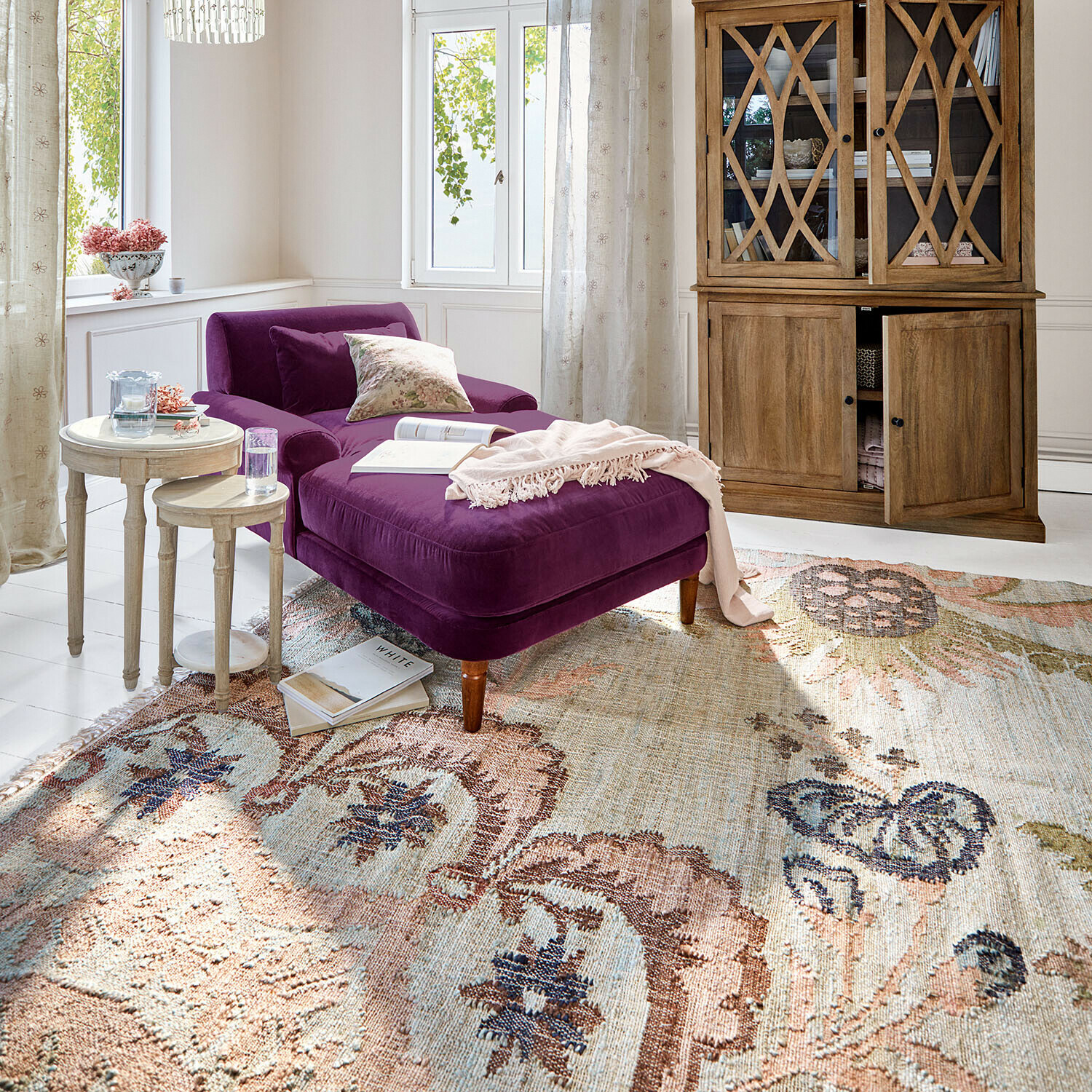 Chaise longue de terciopelo morado en salón luminoso con vitrina de madera y alfombra decorativa