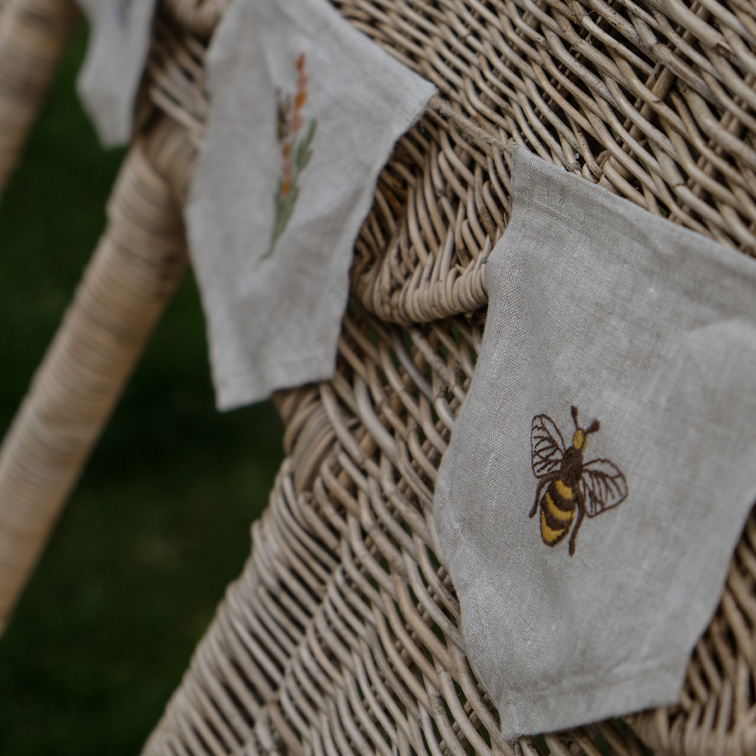 Bolsas decorativas de lino con bordado de abeja y flores sobre silla de mimbre beige
