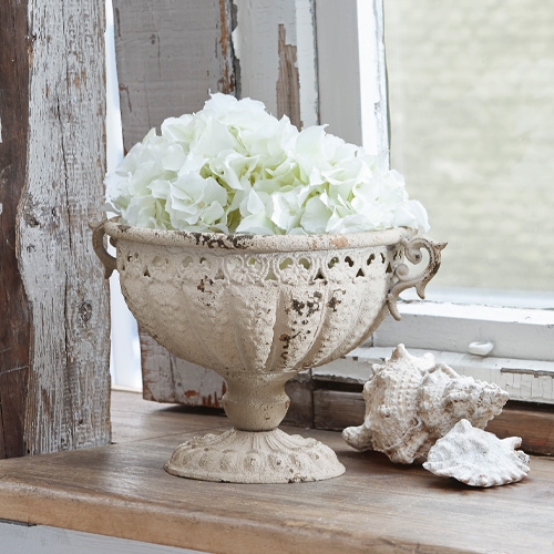 Ánfora decorativa Vescemont de metal crema con flores blancas en alféizar de madera