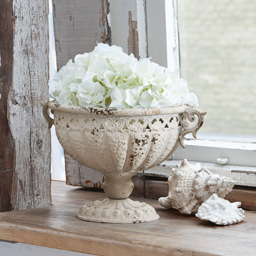 Ánfora decorativa Vescemont de metal crema con flores blancas en alféizar de madera