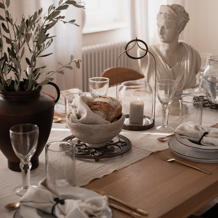 Mesa rústica con cuenco de piedra para pan, vela bajo campana de cristal y escultura blanca decorativa