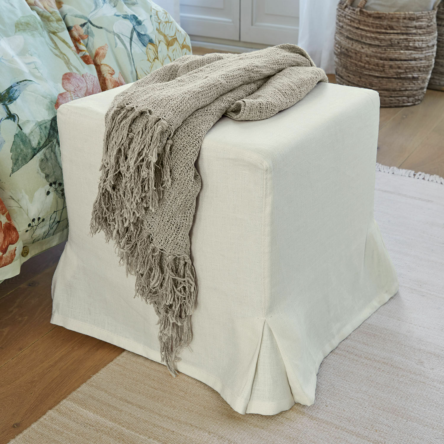 Puf beige de tela con manta tejida gris sobre suelo de madera en interior acogedor