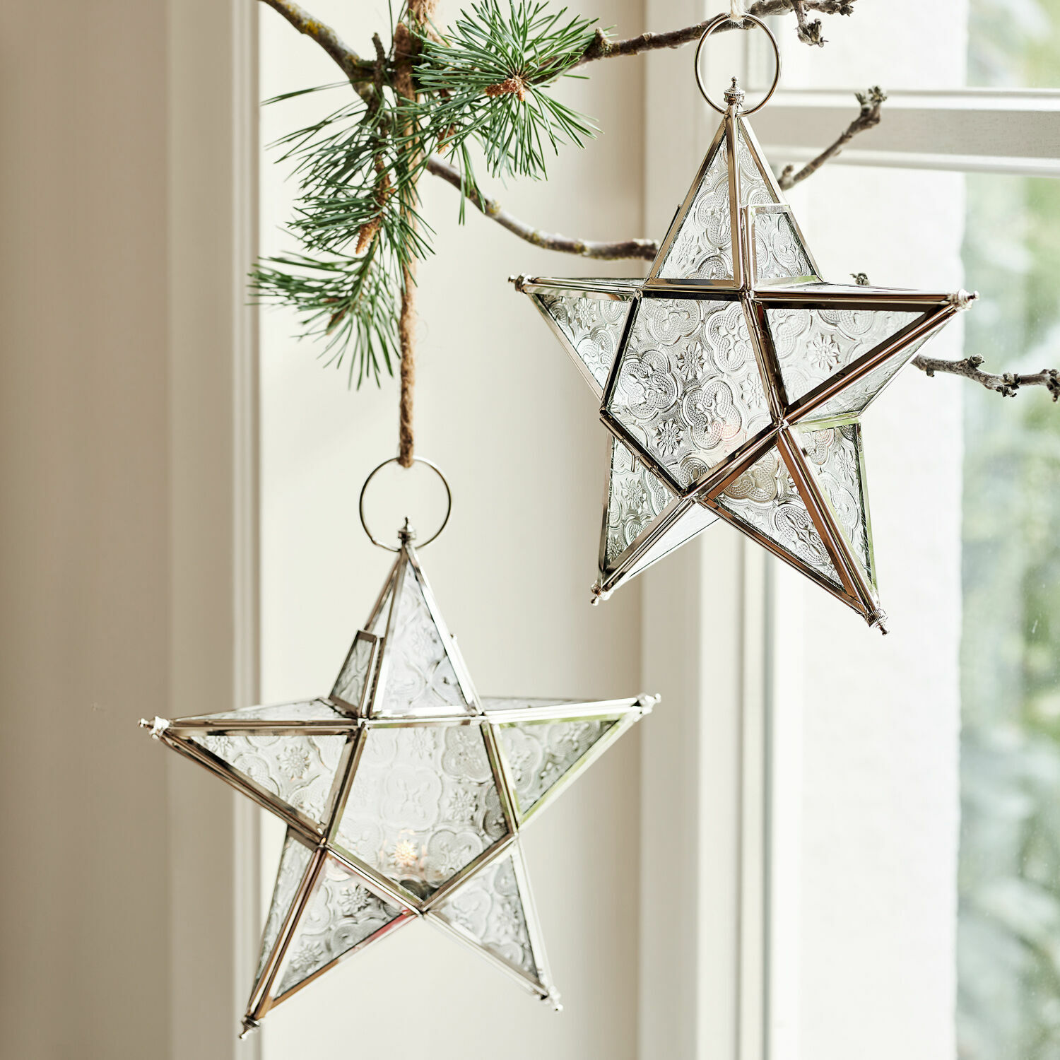 Set de 2 farolillos Kian Estrellas decorativas de vidrio con marco metálico plateado colgadas de una rama de pino