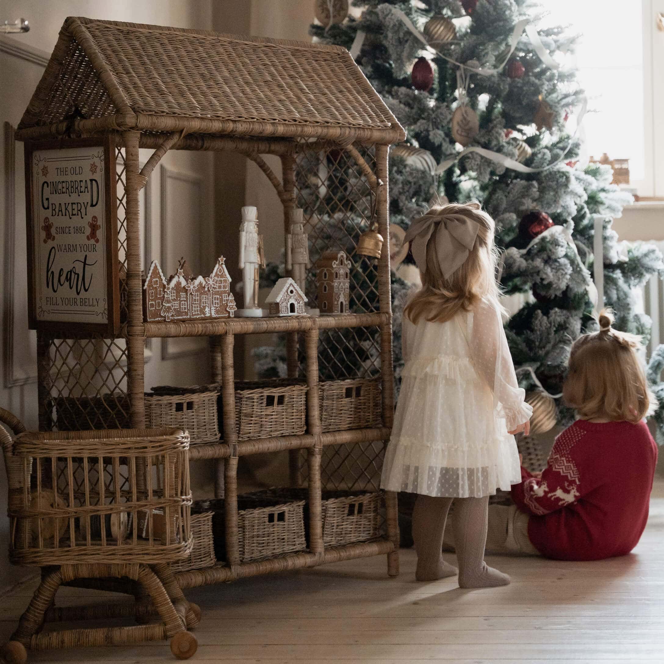 Casa de mimbre con decoraciones de pan de jengibre junto a niños frente a árbol de Navidad decorado