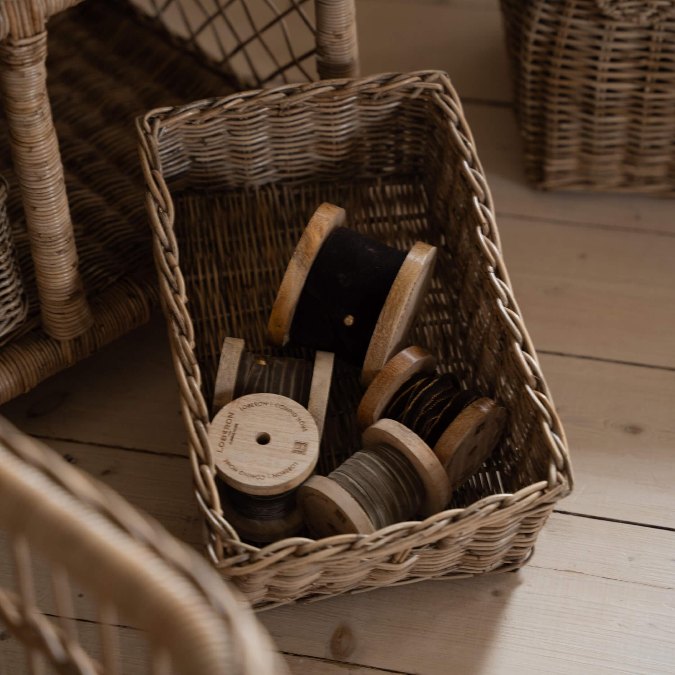 Cesta de mimbre con carretes de madera con hilo negro y gris sobre suelo de madera clara