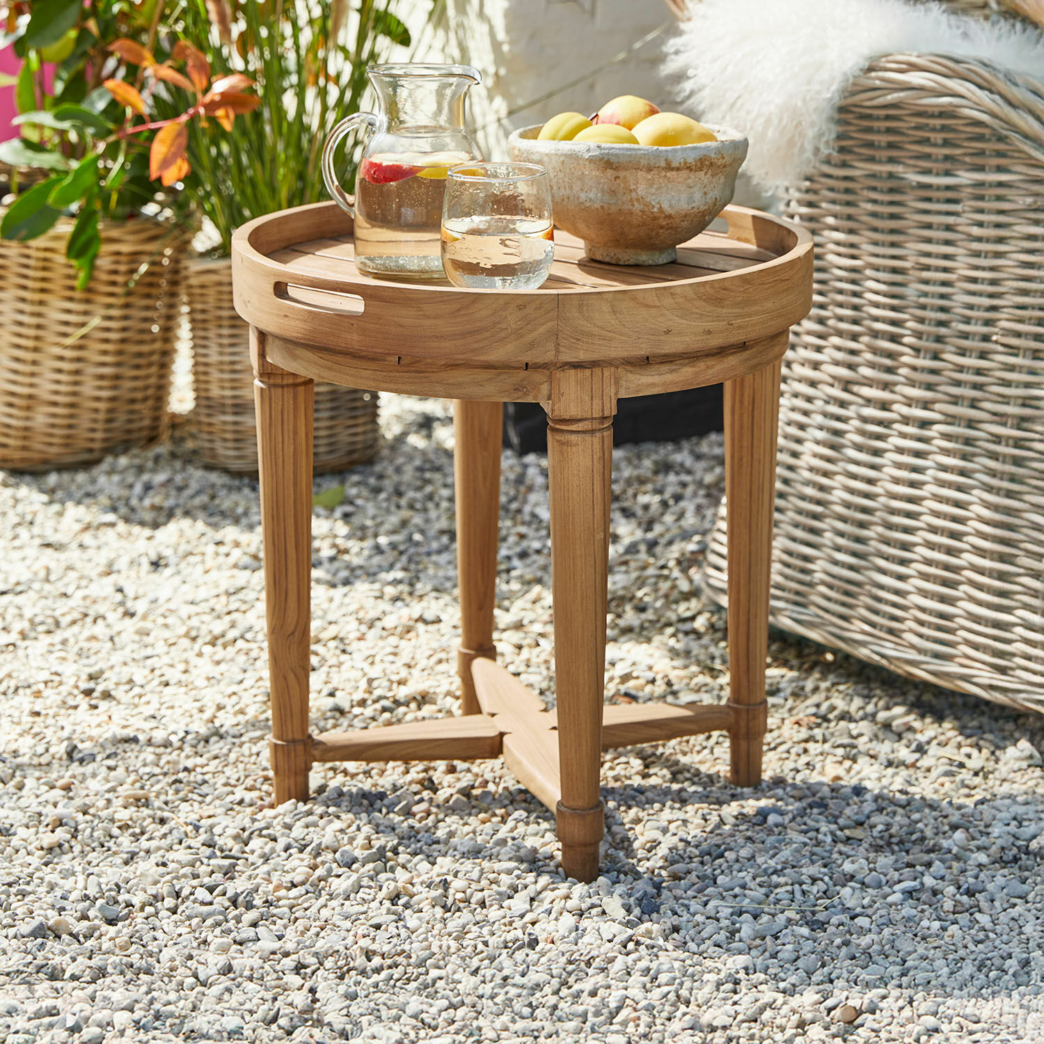 Mesa redonda de madera de teca natural con jarra y cuenco de frutas en exterior sobre grava