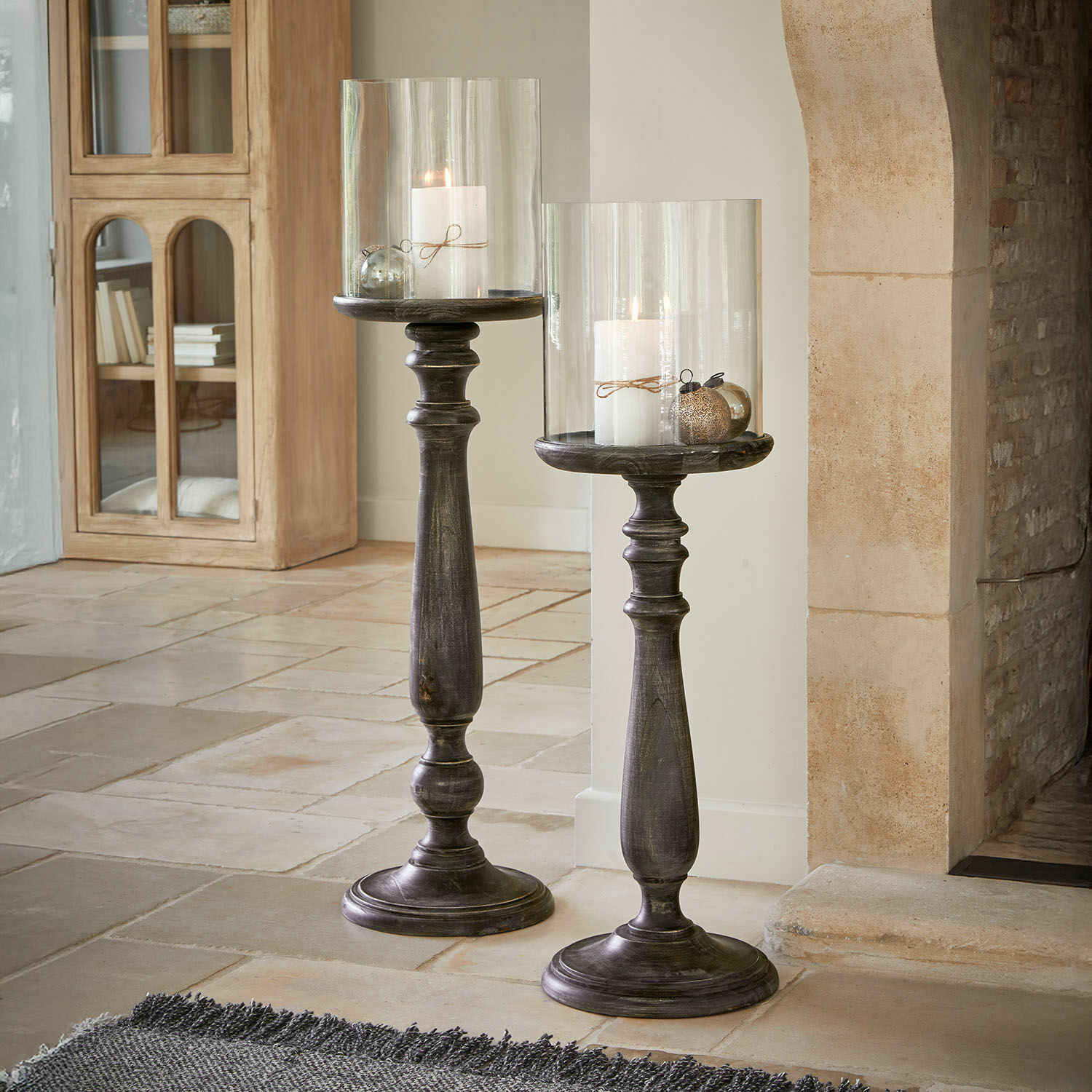 Set de 2 candeleros Reville Dos candelabros altos de madera oscura con pantallas de vidrio transparente y velas blancas en sala de estar