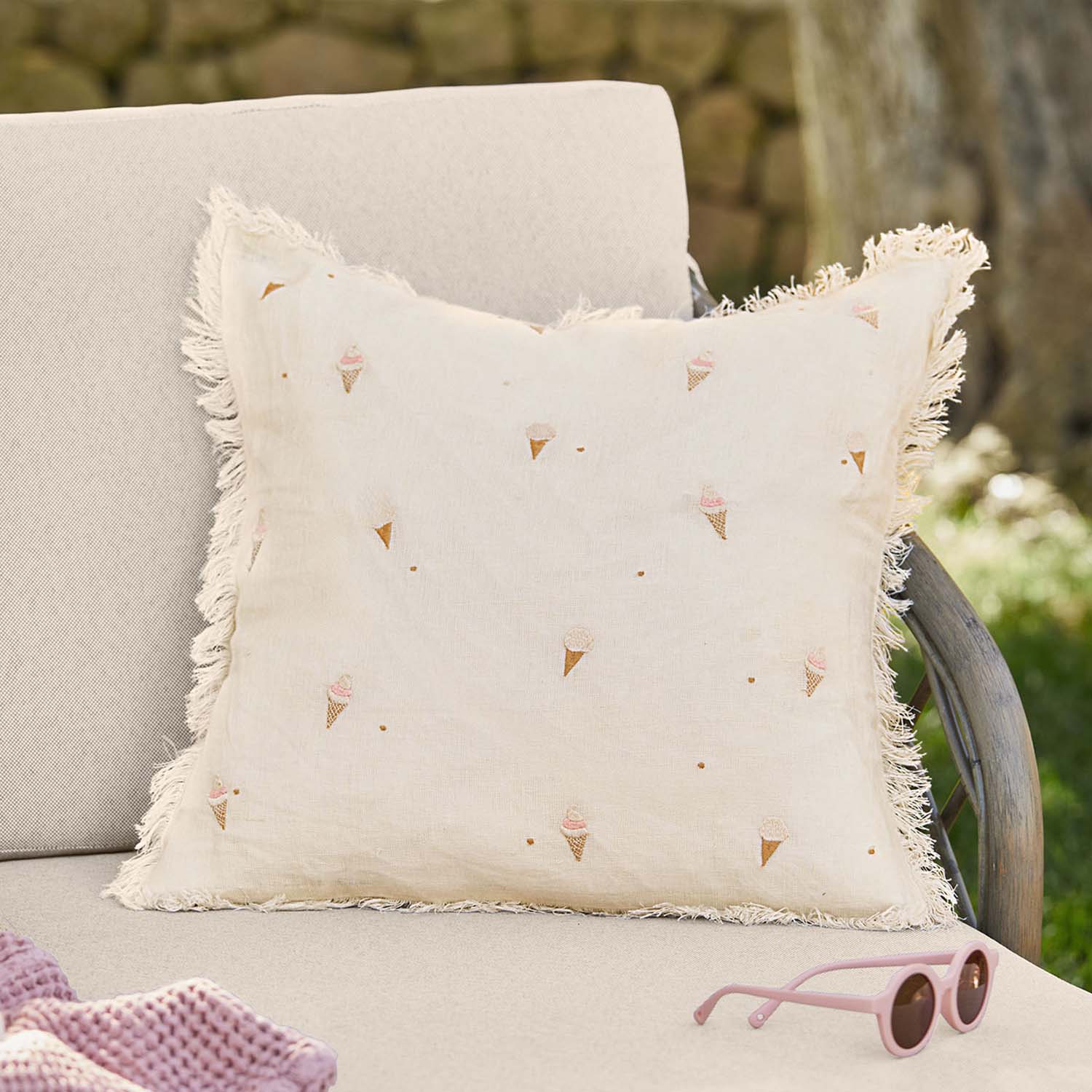 Cojín de lino beige con flecos y estampado de helados en tonos pastel sobre sofá de jardín