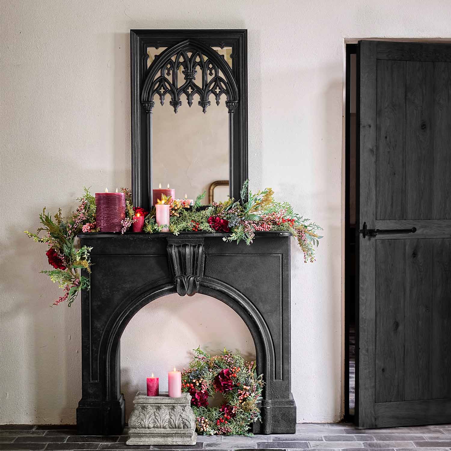 Chimenea negra de piedra con espejo gótico, velas burdeos y rosas rodeadas de decoración vegetal verde