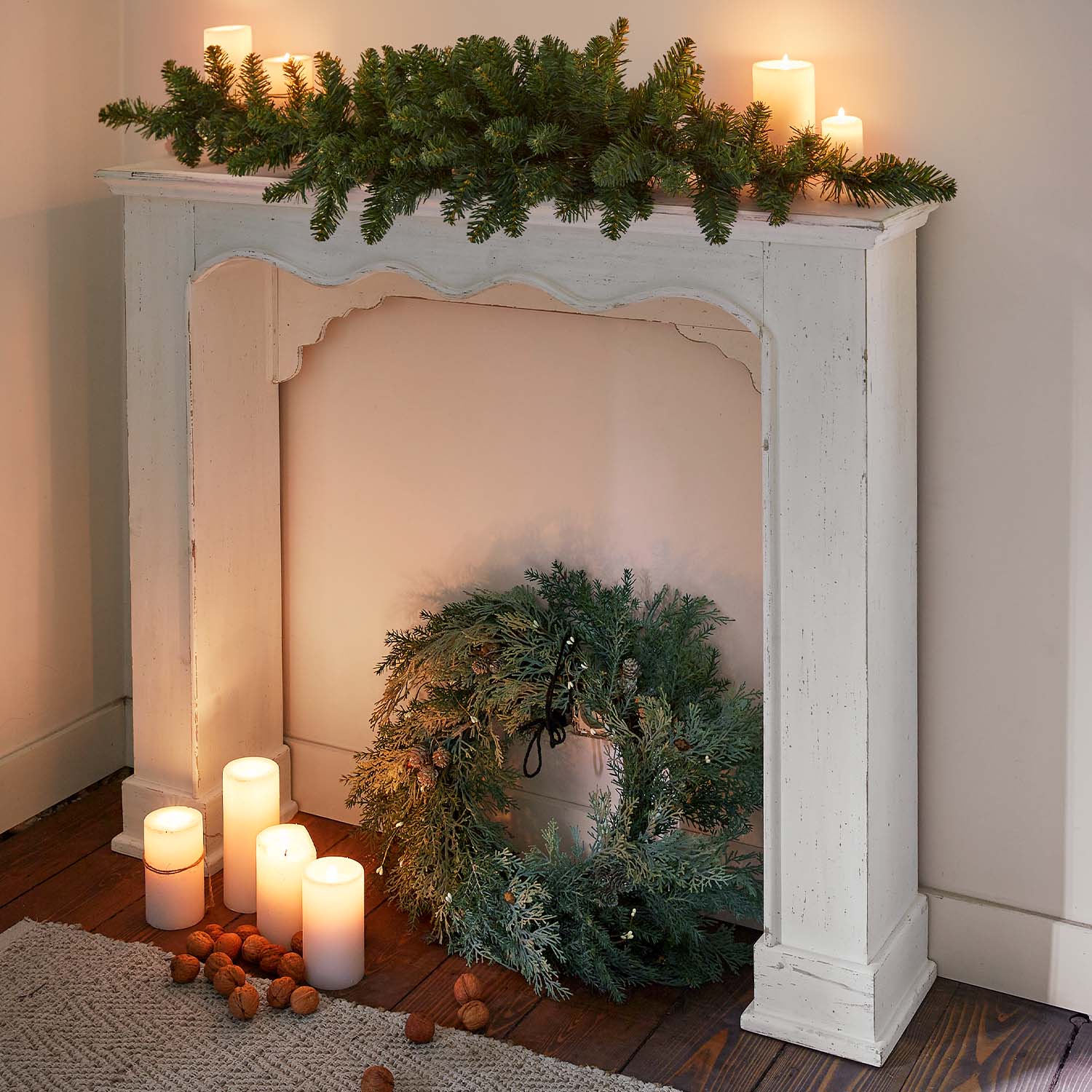 Marco de chimenea de madera blanca con corona verde y velas encendidas sobre suelo de madera, decoración navideña