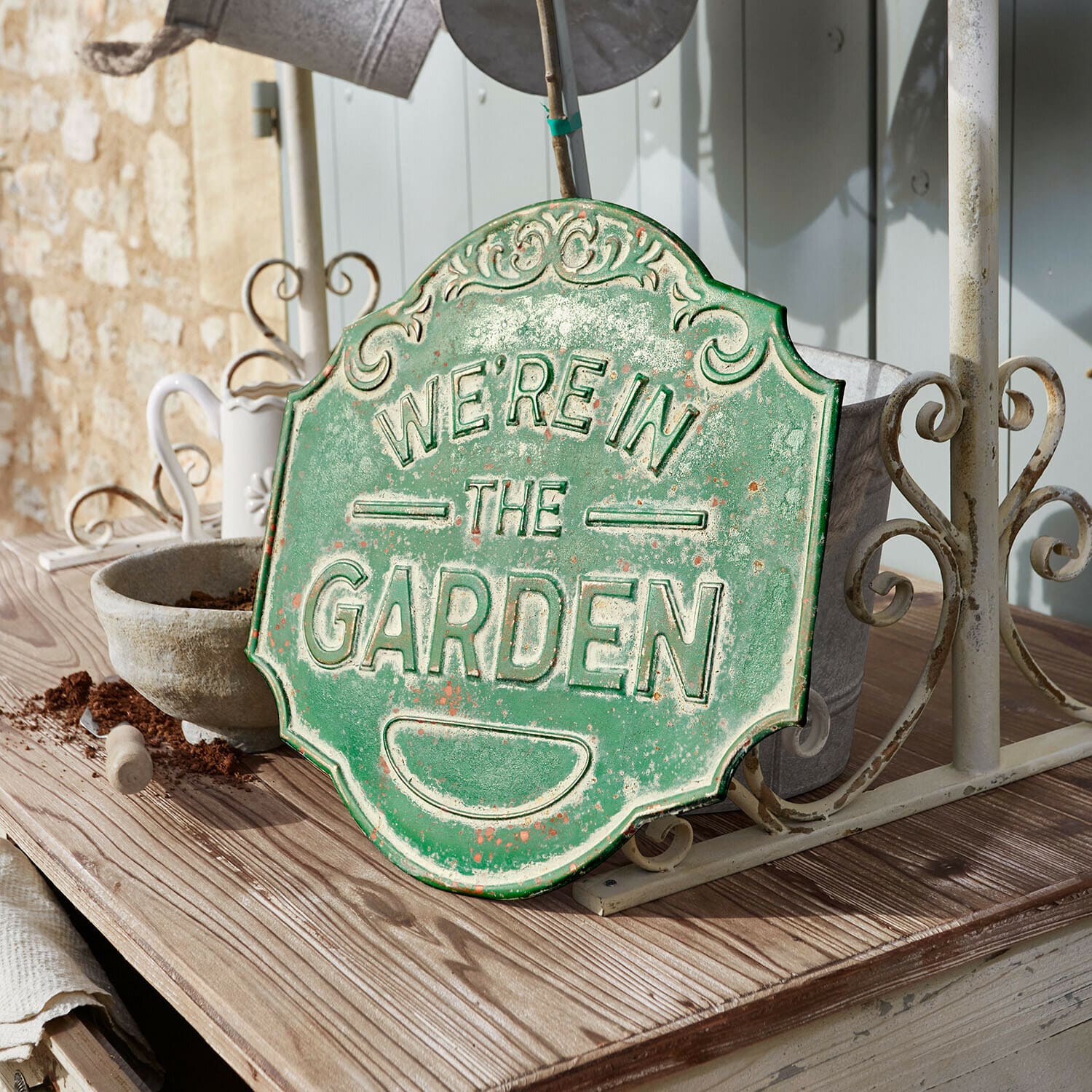 Tablero decorativo Merrin Cartel decorativo de metal verde con texto We're in the Garden sobre mesa de madera en jardín