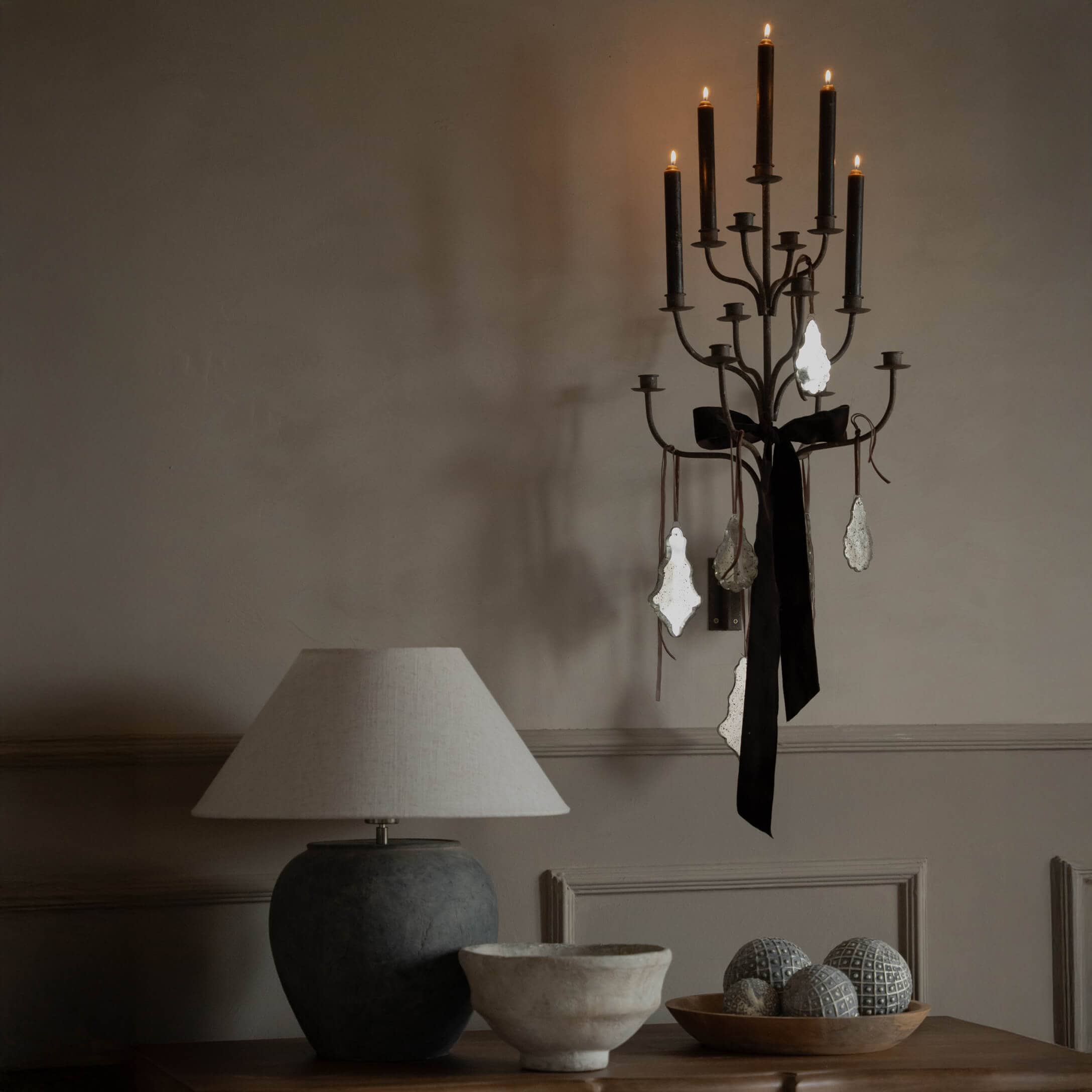 Candelabro de pared negro con lazo negro y adornos, lámpara beige sobre mesa de madera con decoración