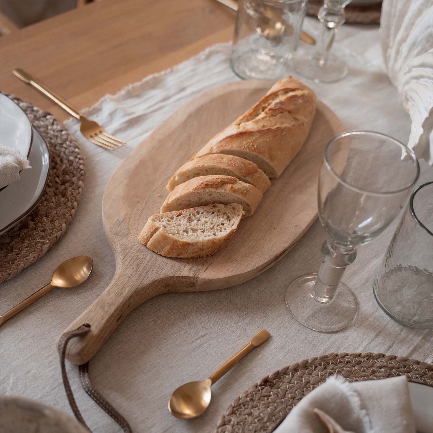 Tabla de cortar de madera con baguette cortada sobre mesa con mantel natural y cubiertos dorados