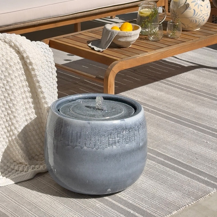 Fuente de mesa de cerámica gris sobre alfombra clara junto a mesa de madera y manta blanca