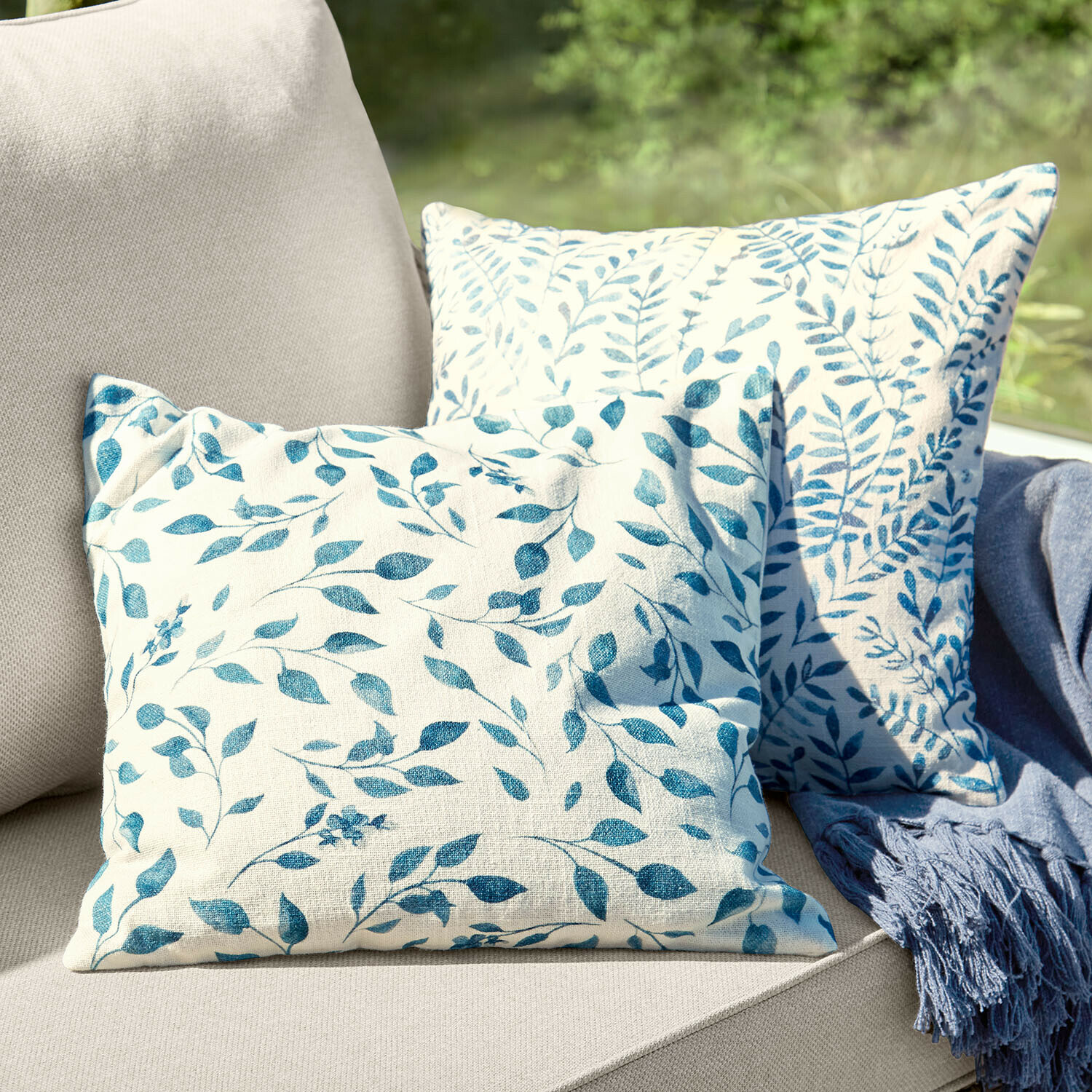 Set de 2 fundas de cojín Ocevara Dos cojines decorativos de algodón crema con estampado de hojas azules sobre sofá beige