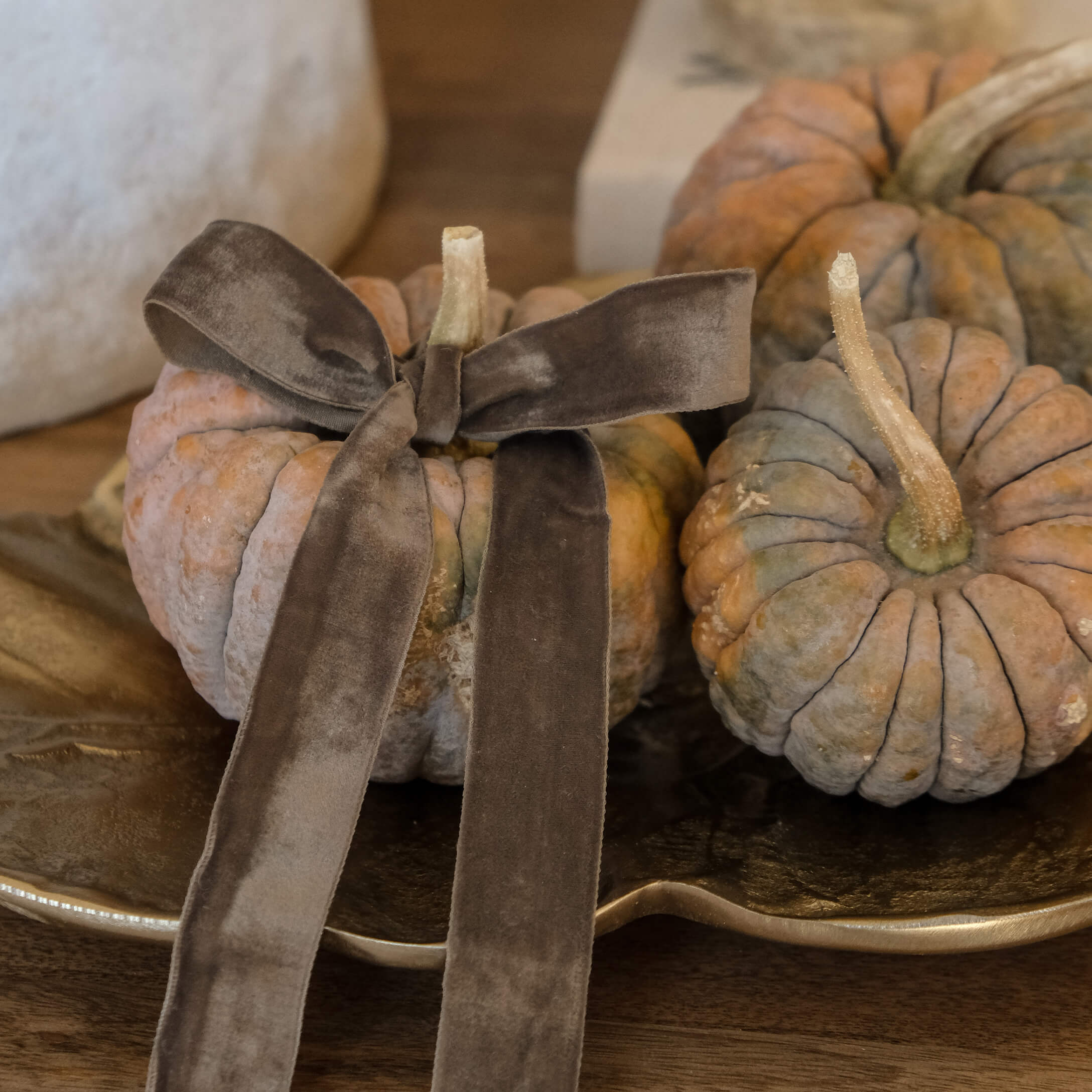 Calabazas decorativas naranja-gris con lazo de terciopelo marrón sobre bandeja dorada en mesa de madera