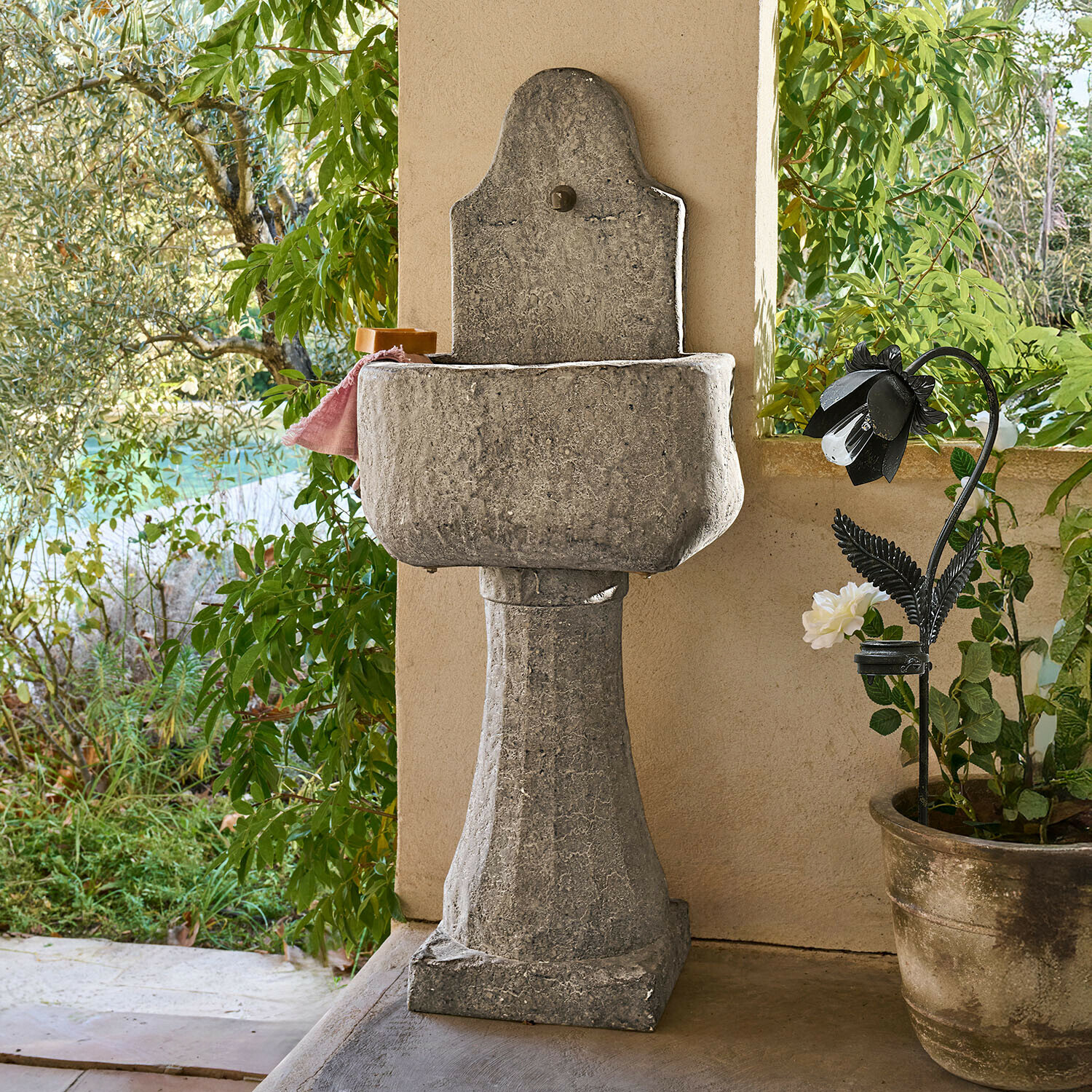 Lavabo de jardín de piedra gris sobre columna con acabado natural y plantas verdes alrededor