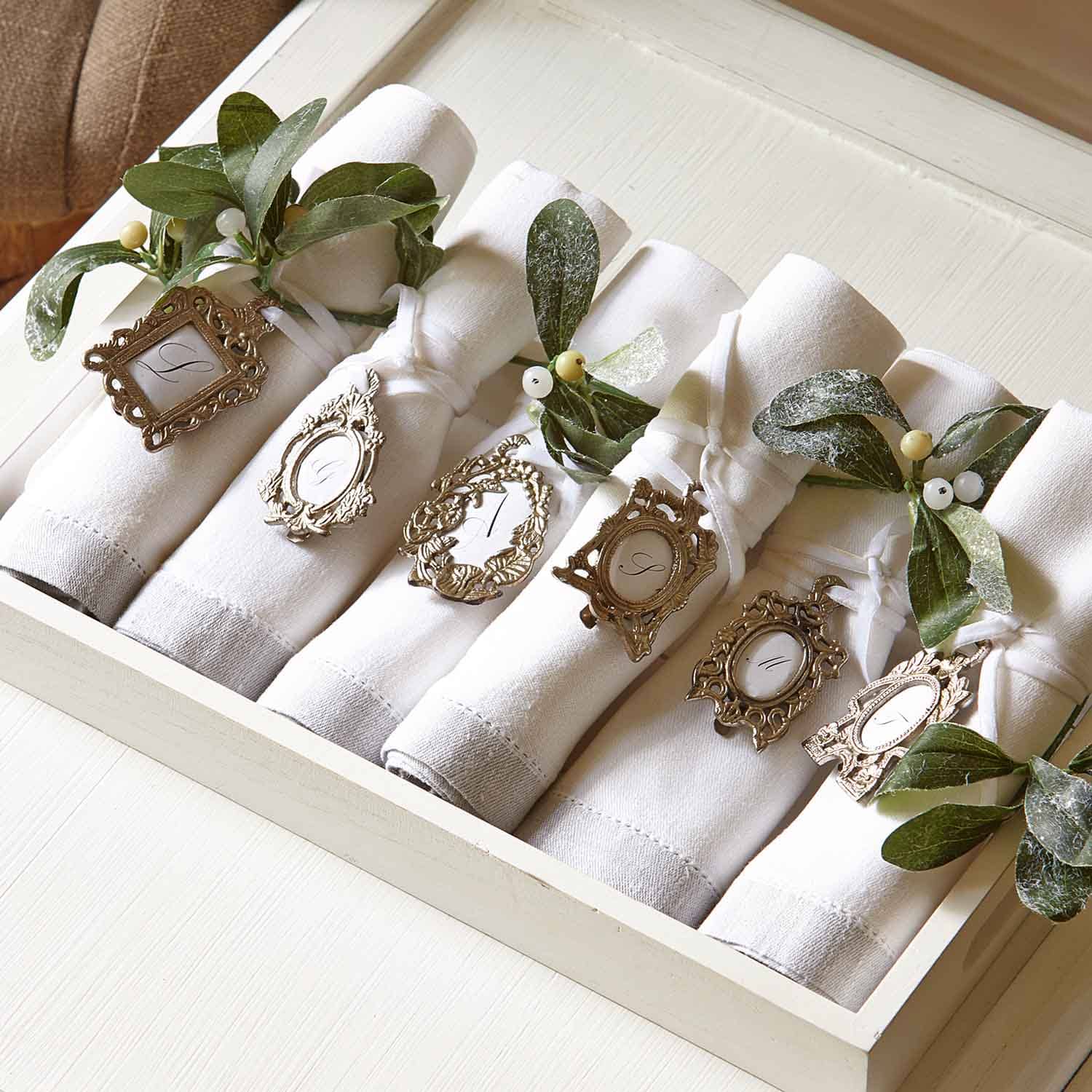 Servilletas de lino blancas con anillos metálicos decorativos y hojas verdes para mesa elegante.