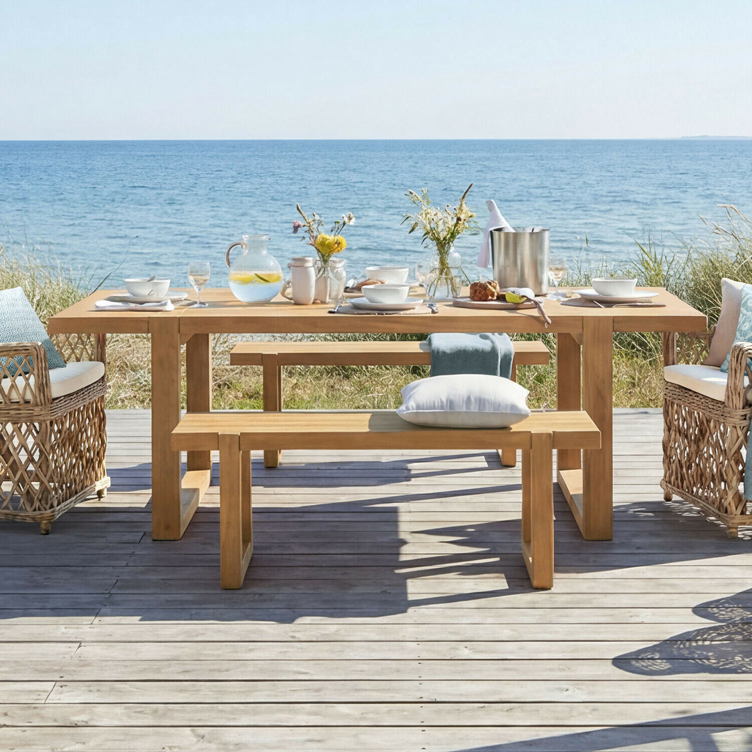 Mesa Halinor Mesa de jardín de madera con bancos y sillones de mimbre frente al mar, con vajilla y flores