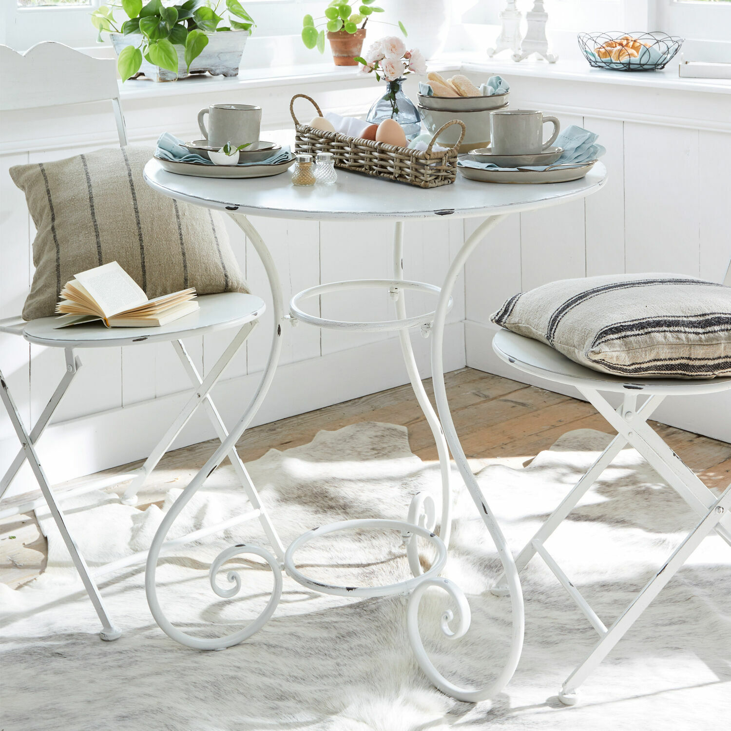 Mesa Iteuil Mesa metálica blanca con dos sillas plegables y cojines decorativos en interior luminoso