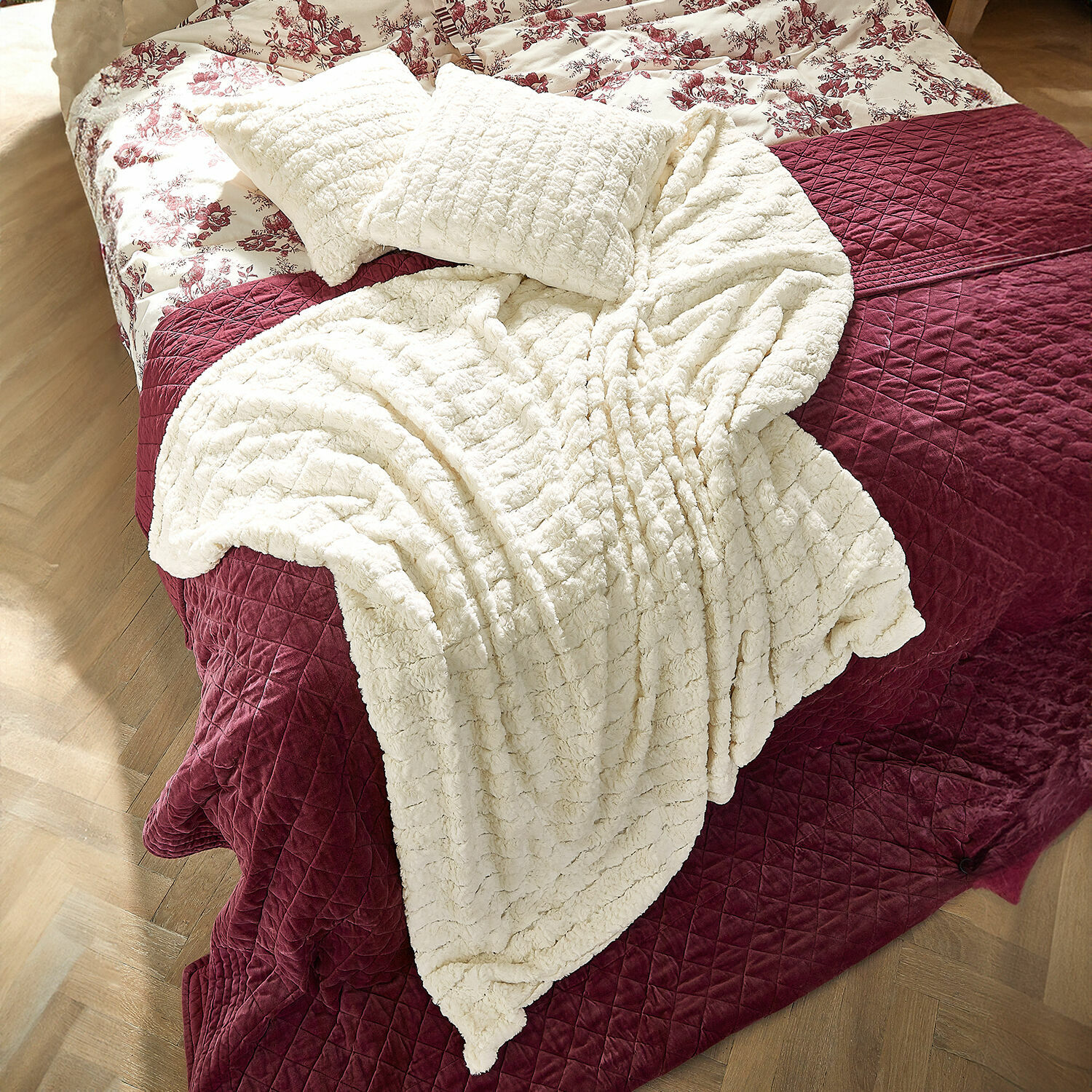 Plaid Dorelice Manta y cojines de felpa crema sobre colcha acolchada burdeos en la cama