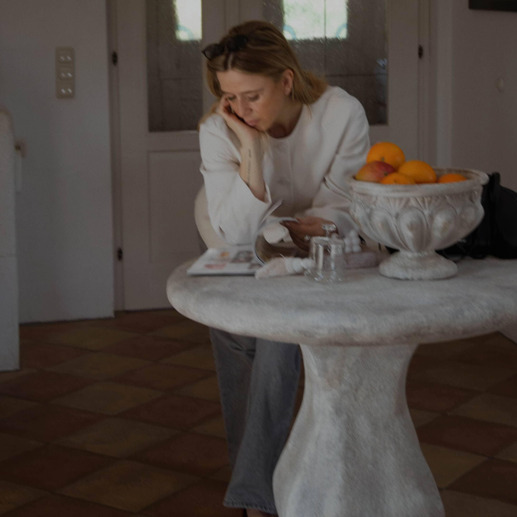 Mujer con suéter blanco leyendo junto a una mesa redonda de piedra con frutero en casa