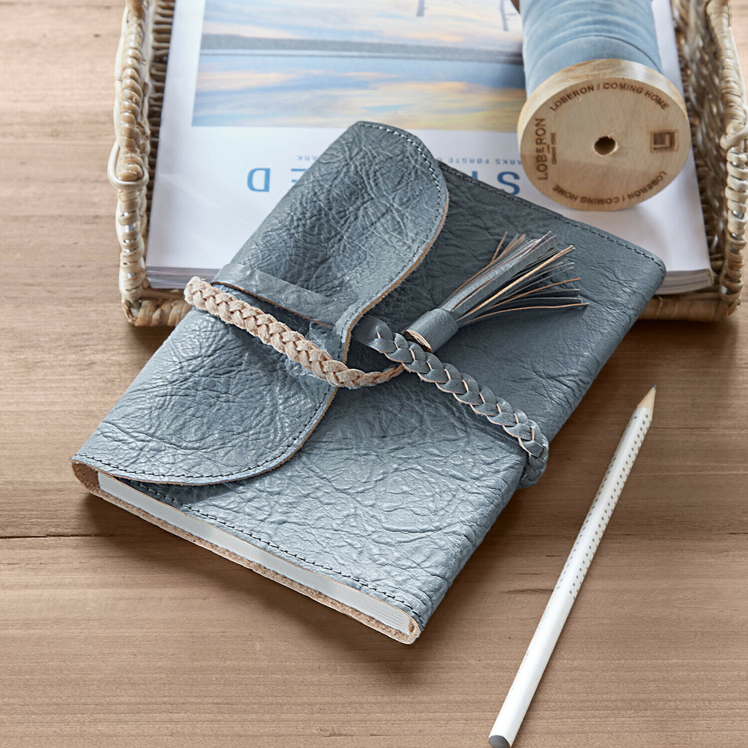 Cuaderno de cuero gris con cierre trenzado y borla sobre mesa de madera con lápiz blanco