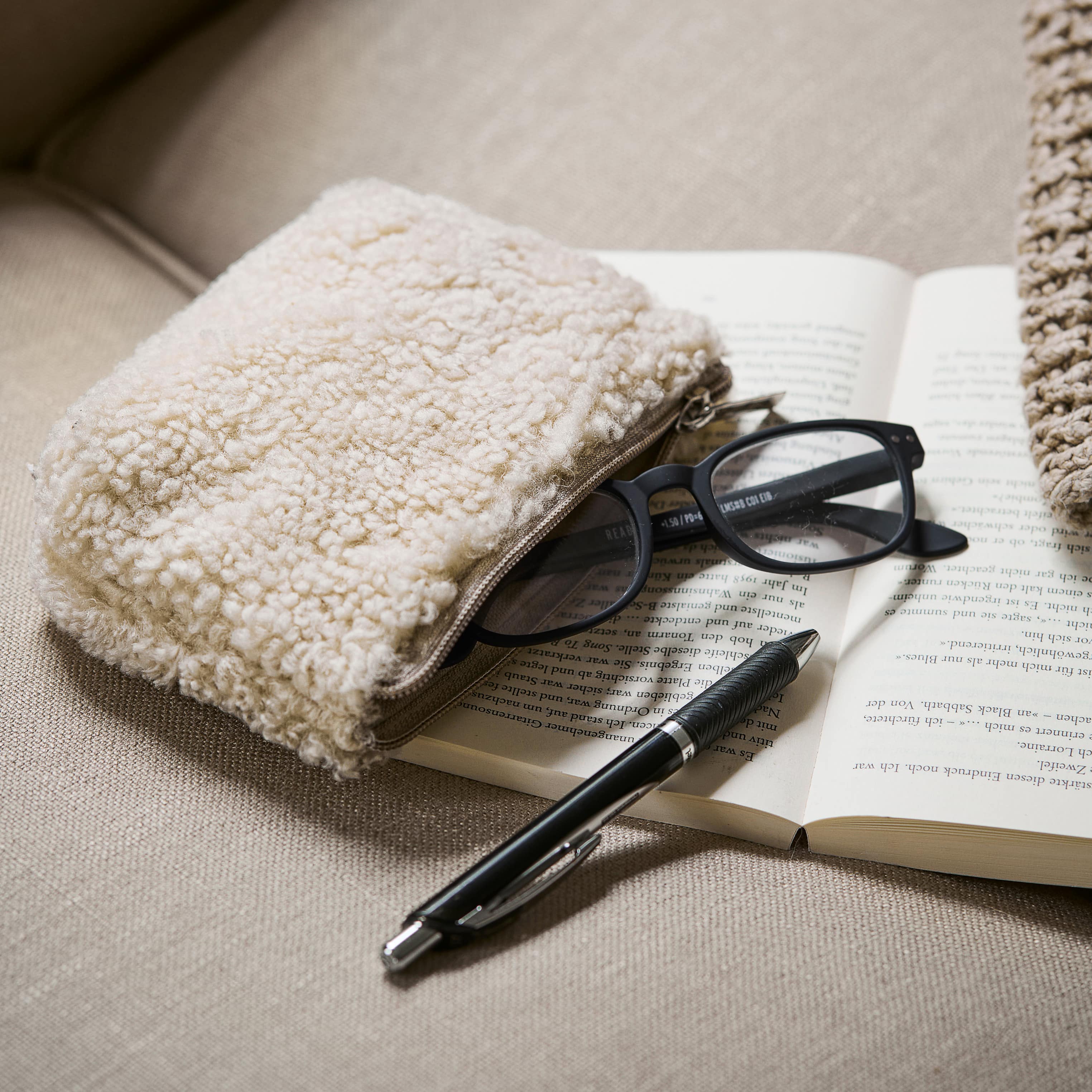 Neceser beige de tela suave sobre sofá junto a gafas y bolígrafo sobre libro abierto