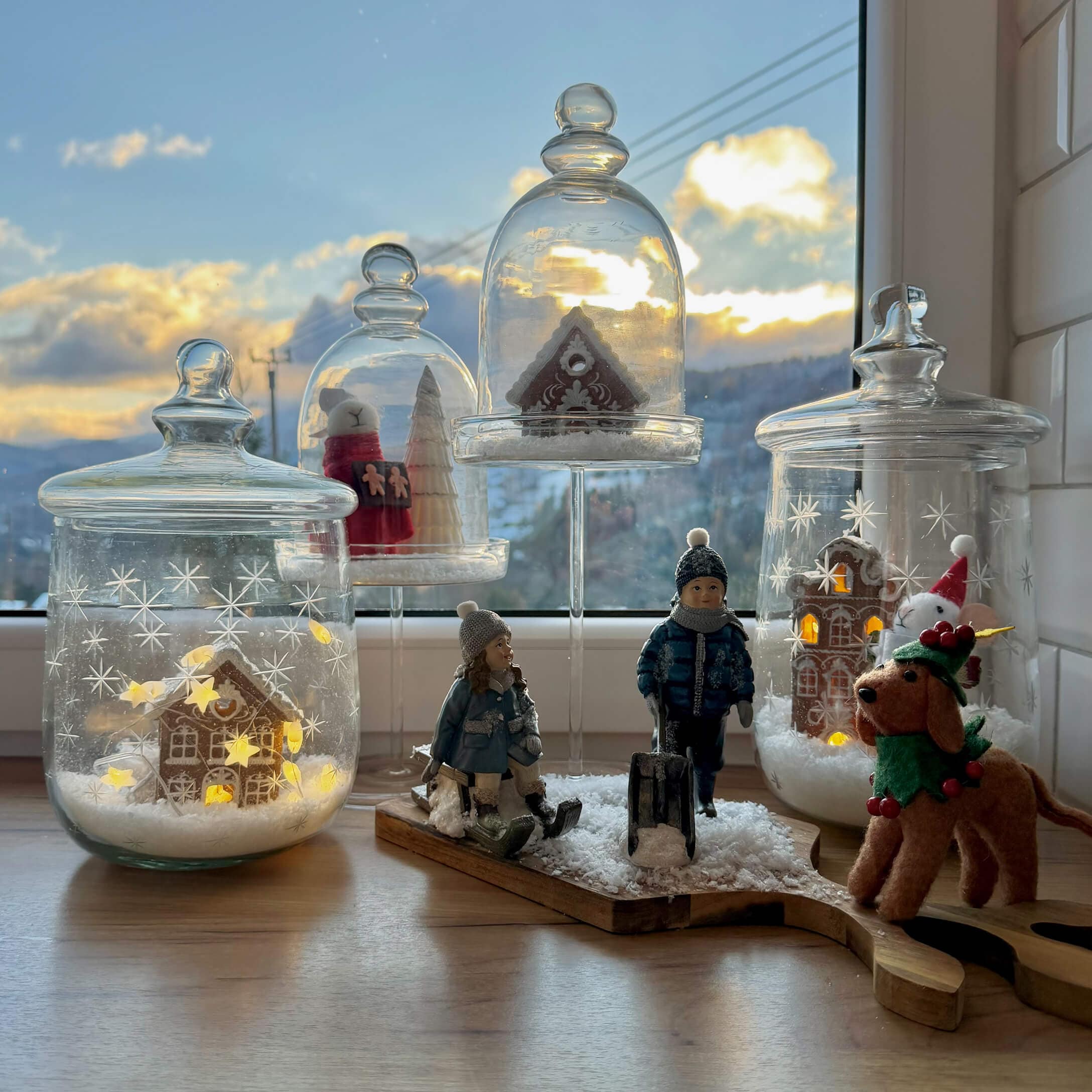 Decoración navideña con figuras de niños, perro y casas de jengibre bajo campanas de cristal en el alféizar