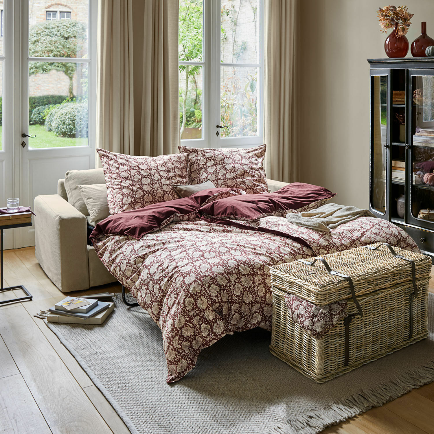 Sofá cama con ropa de cama floral burdeos y crema en sala luminosa con baúl de mimbre