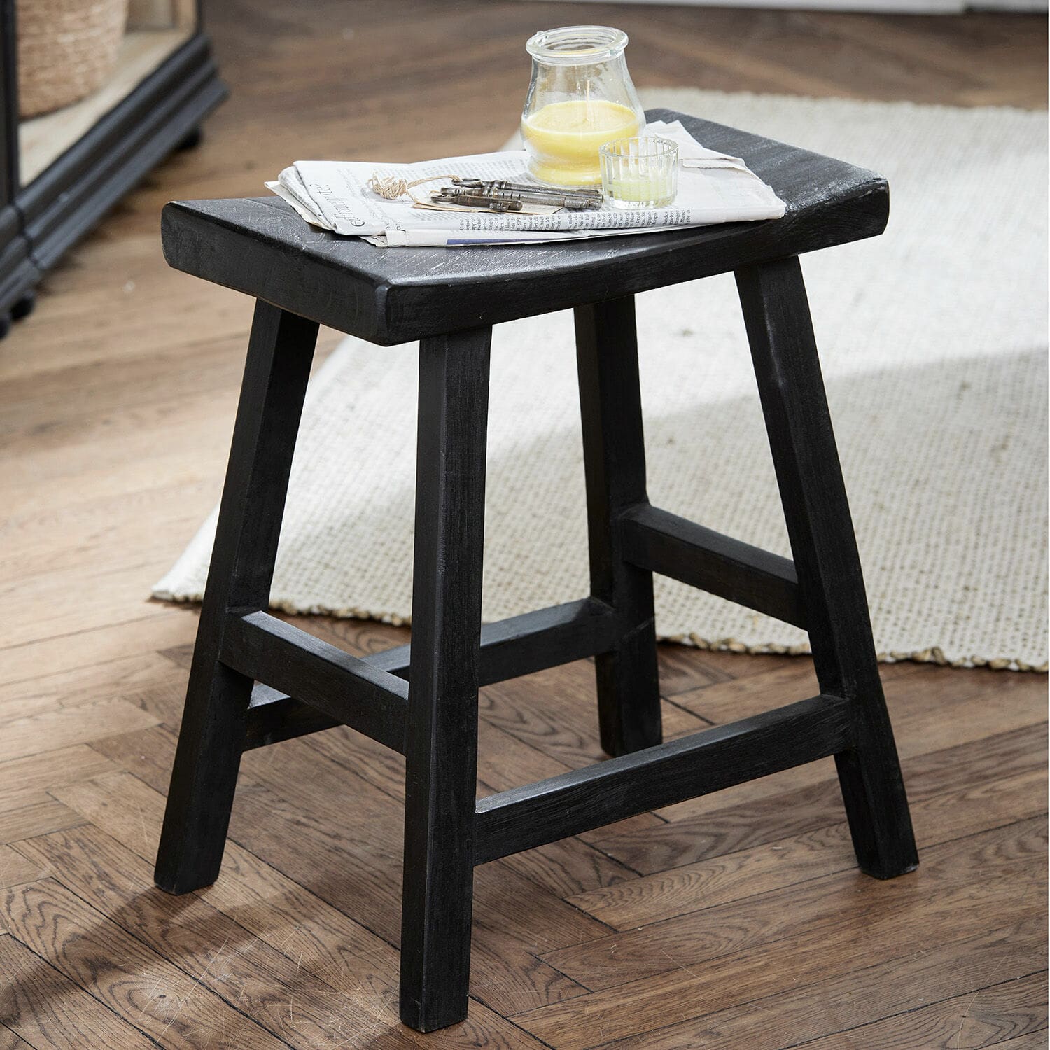 Taburete de madera negro con estructura simple sobre suelo de madera, periódico y velas encima
