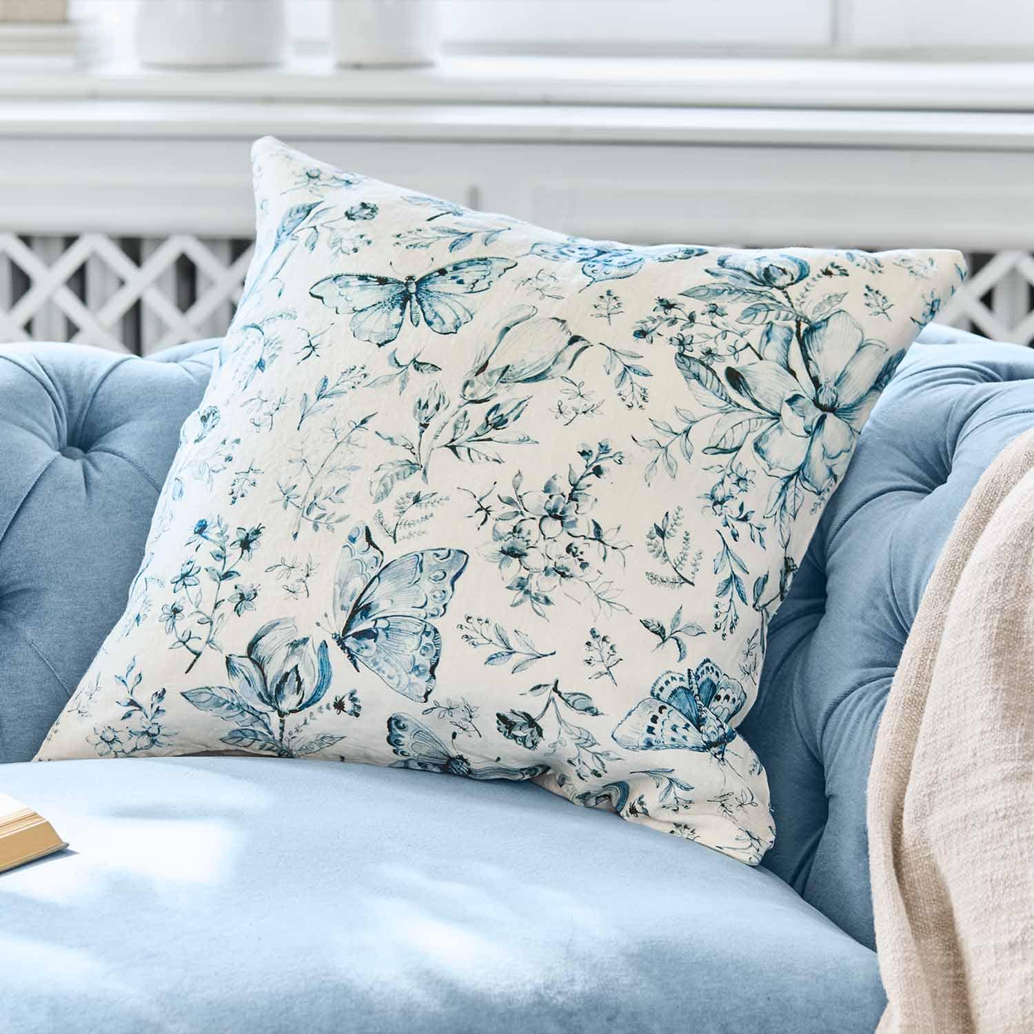 Cojín decorativo de algodón con estampado de mariposas y flores azules sobre fondo claro en sofá azul