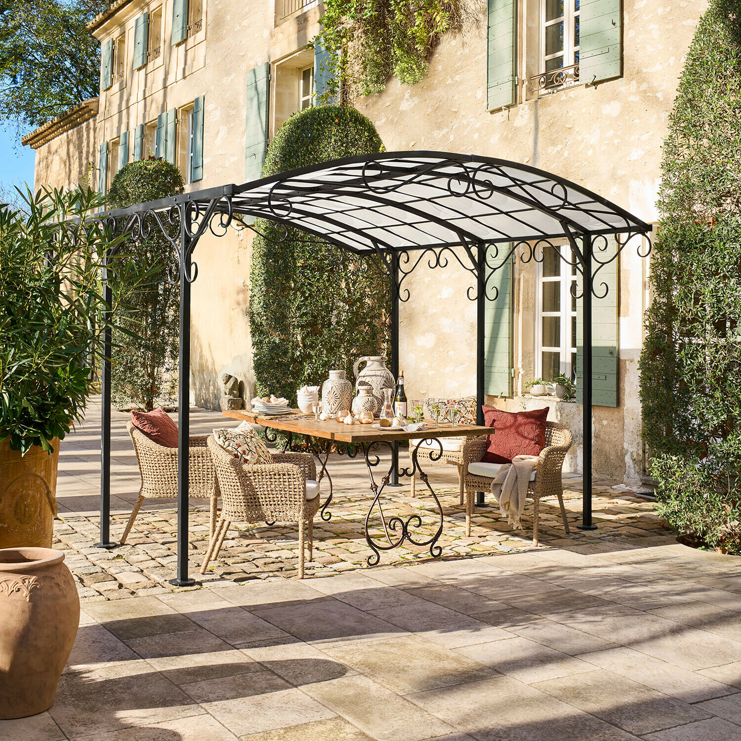 Pérgola Delbridge Pérgola de metal negro con adornos en espiral sobre mesa de madera y sillas de mimbre en patio