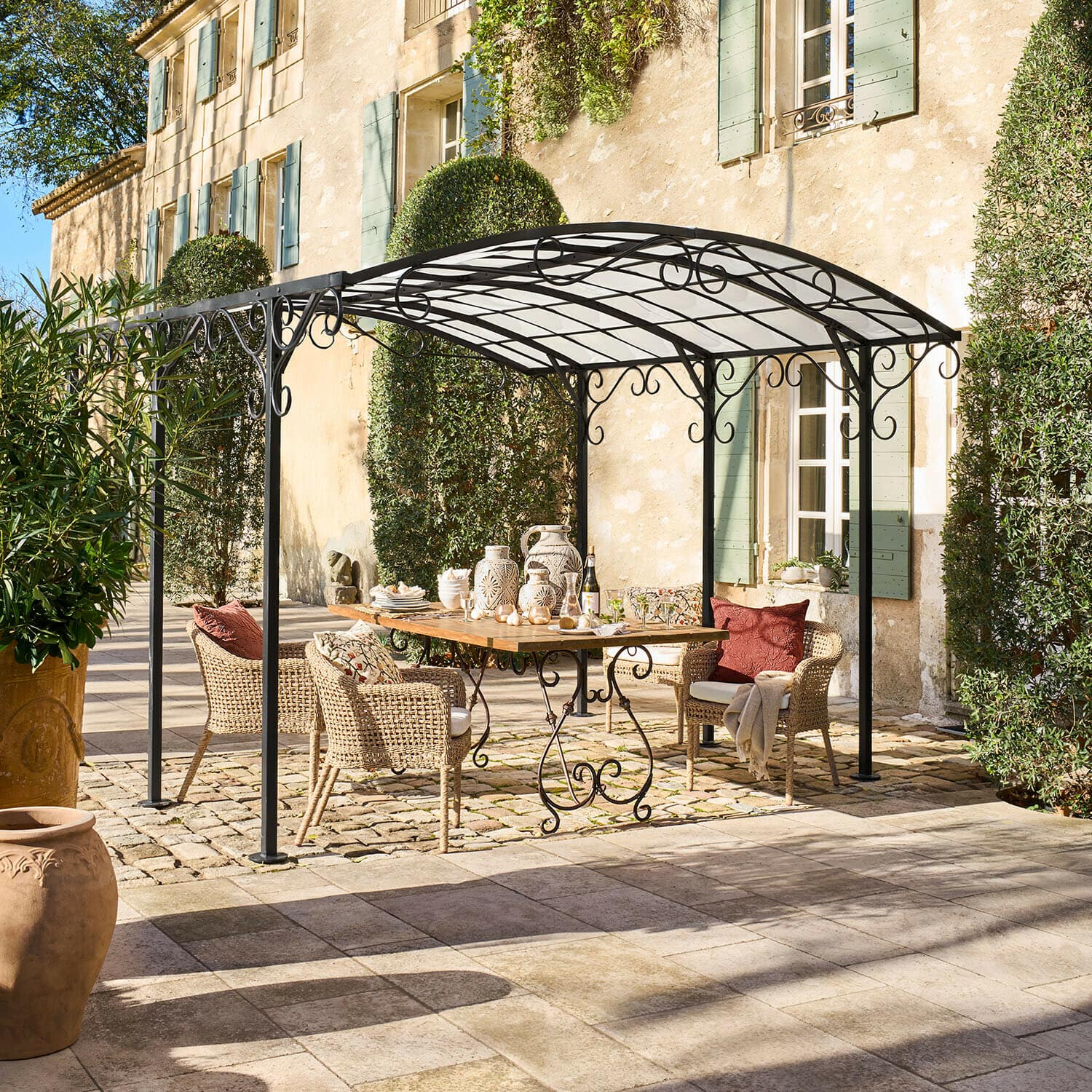 Pérgola Delbridge Pérgola de metal negro con adornos en espiral sobre mesa de madera y sillas de mimbre en patio